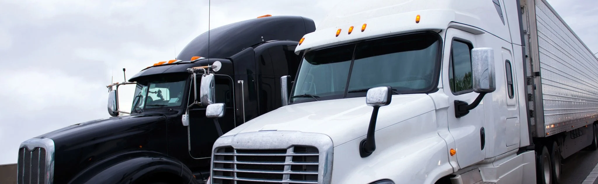 Two semi-trucks, one black and one white, parked side by side under cloudy skies.