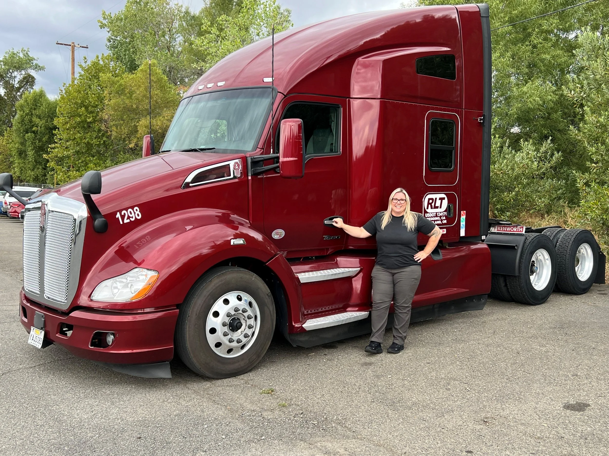 A woman with long blonde hair and glasses standing next to a large red semi-truck, smiling and holding a remote or key fob in her right hand, in a parking lot with trees and cloudy sky in the background.