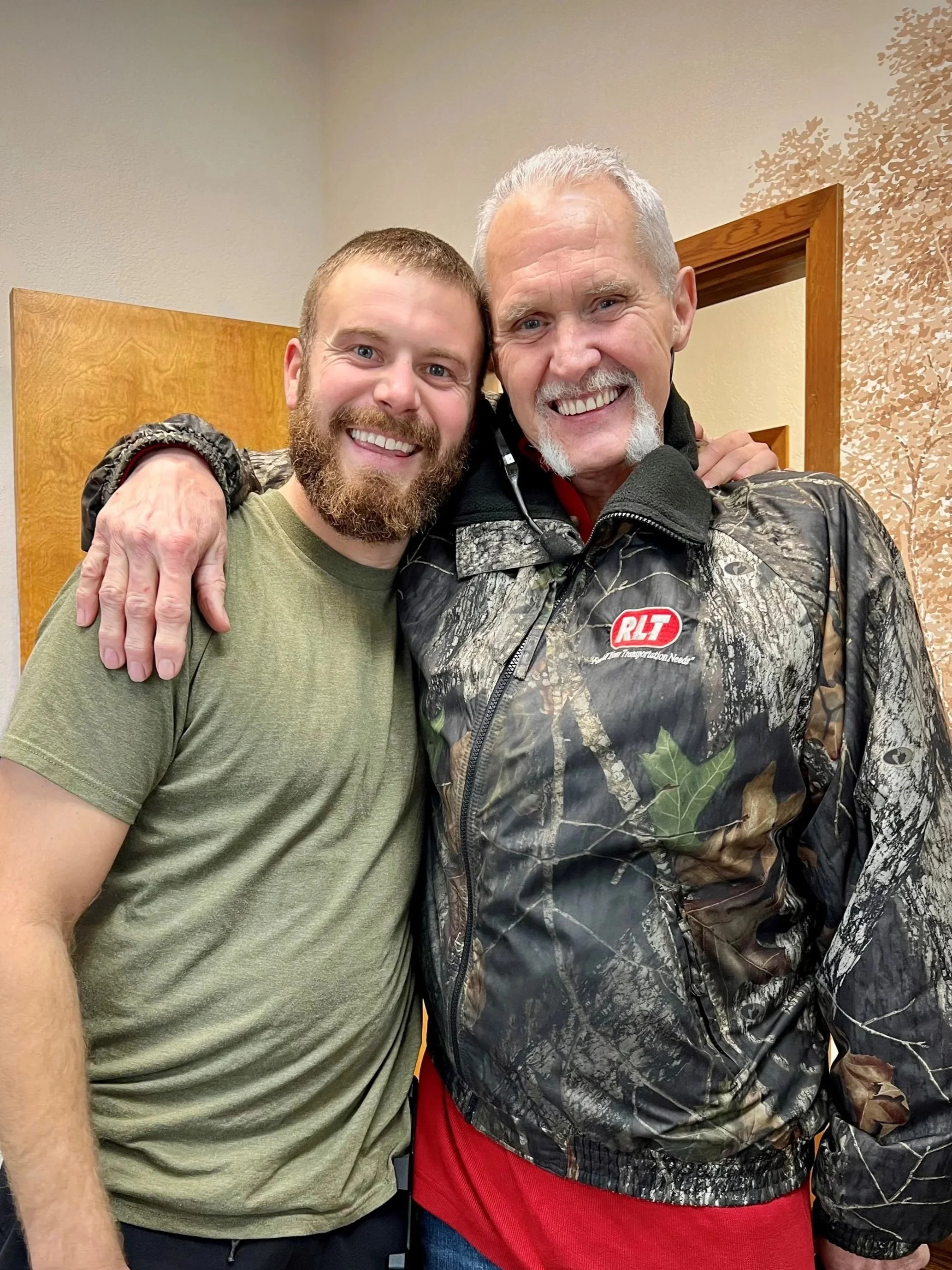Two smiling men, one with a beard and short hair, the other with gray hair and a beard, hugging each other, indoors with a wooden door and beige wall with a tree design in the background.