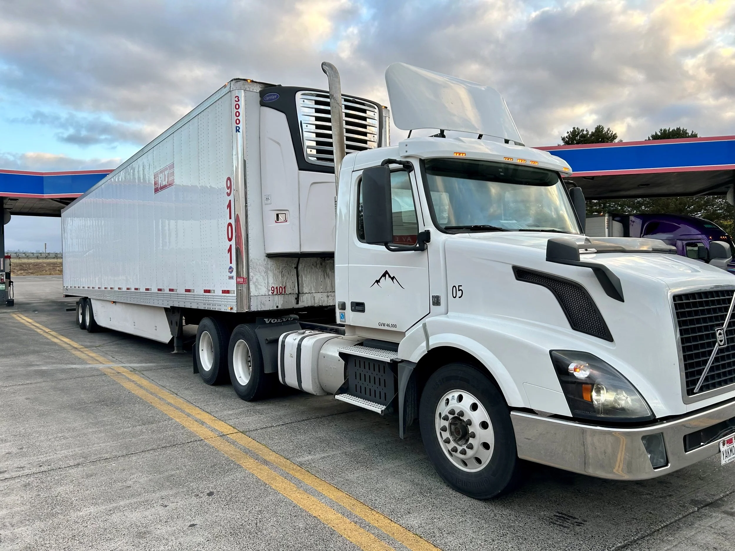 A white semi-truck with a large trailer parked at a gas station or rest stop, with a canopy overhead. The trailer has red and blue markings, and a small mountain logo on the side. The sky is partly cloudy with shades of blue, yellow, and gray.