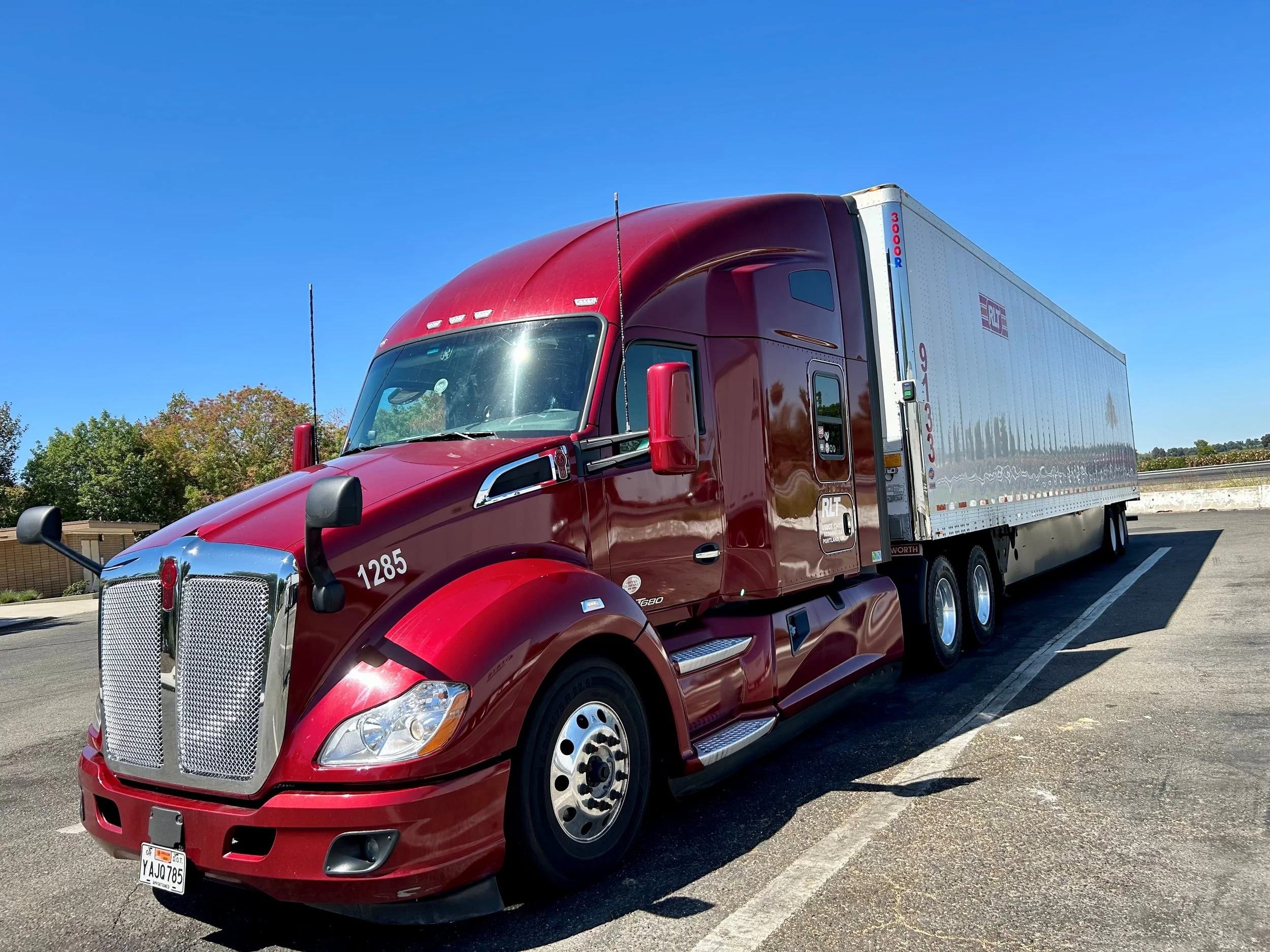 A red semi-truck with a white trailer parked in a parking lot under a clear blue sky, with trees visible in the background.