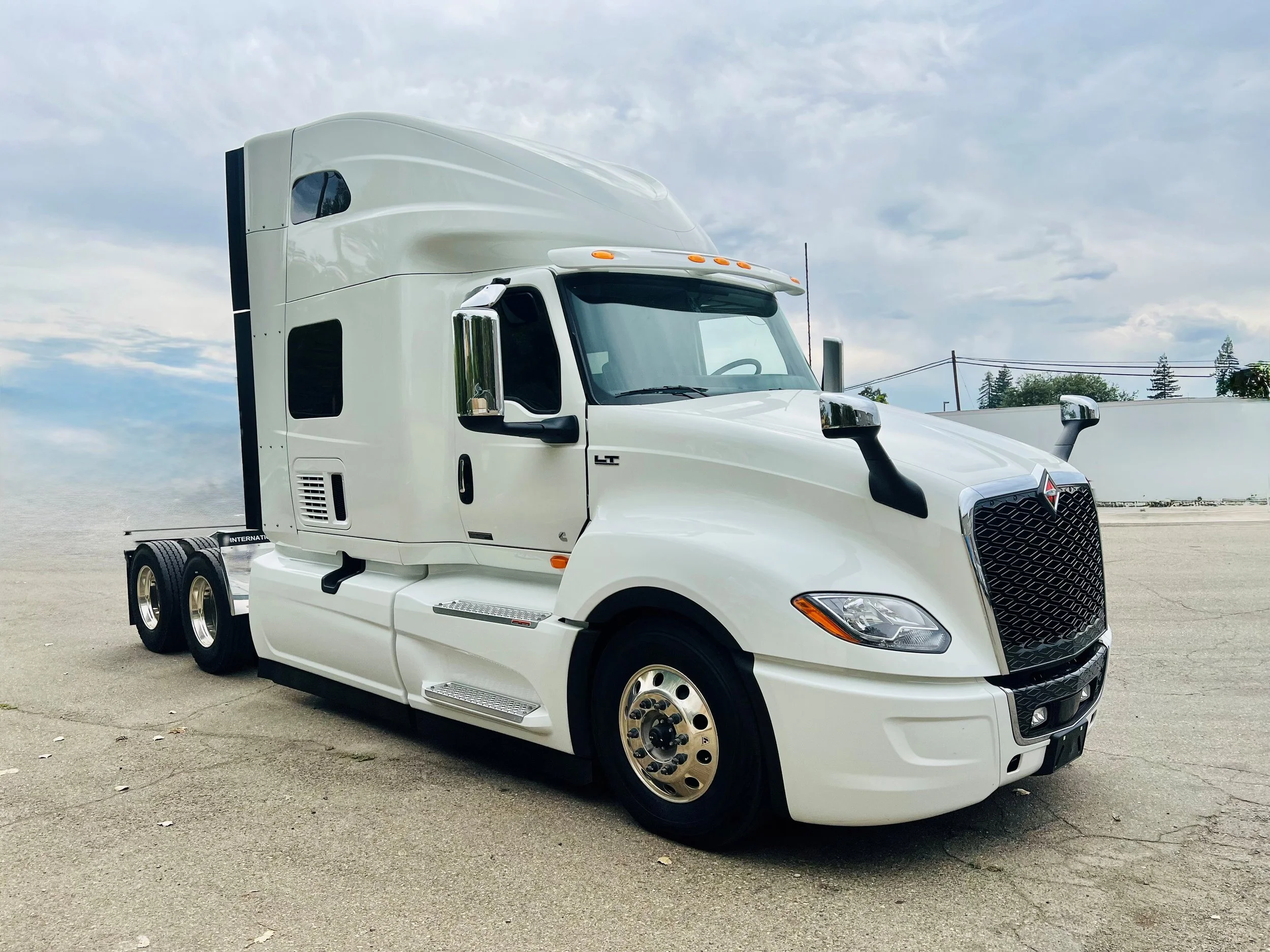 A white semi-truck parked on an empty lot under cloudy skies, with trees and power lines in the background.