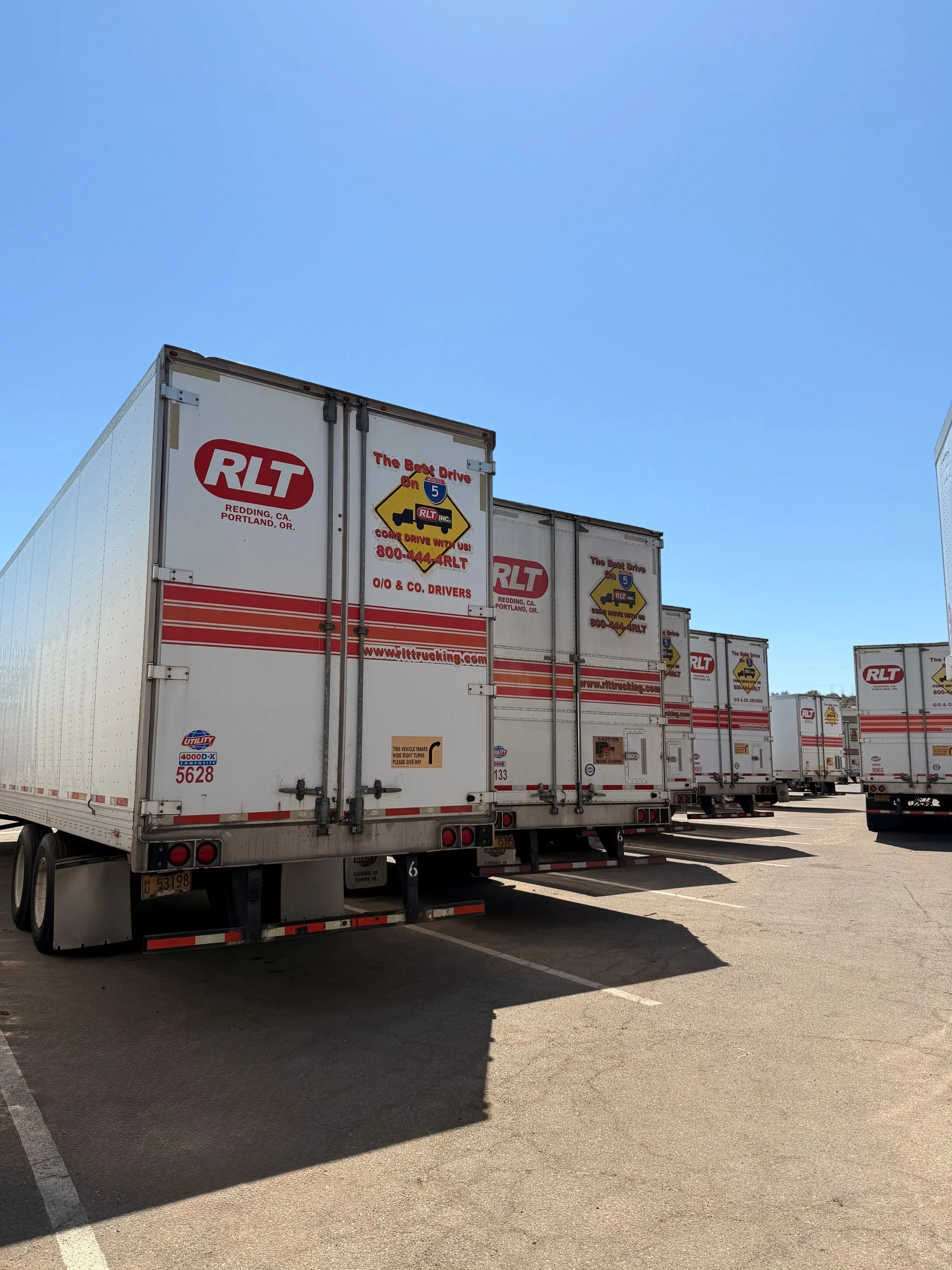 A row of white delivery trucks with RLT logos parked in a lot under a clear blue sky.