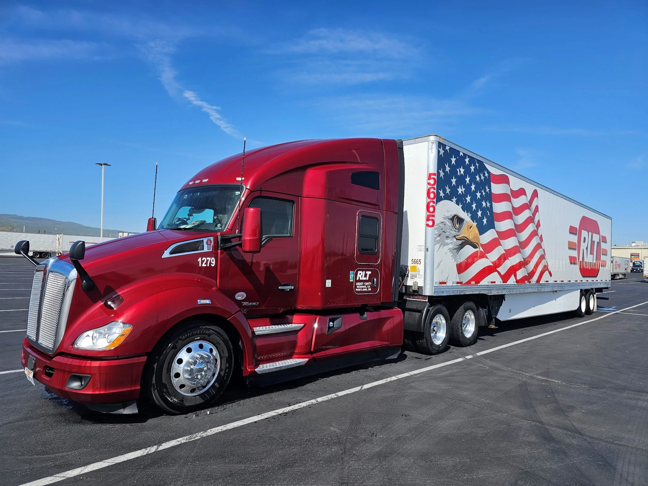A red semi-truck with a white trailer decorated with an American flag and an eagle, parked in a lot under a clear blue sky.