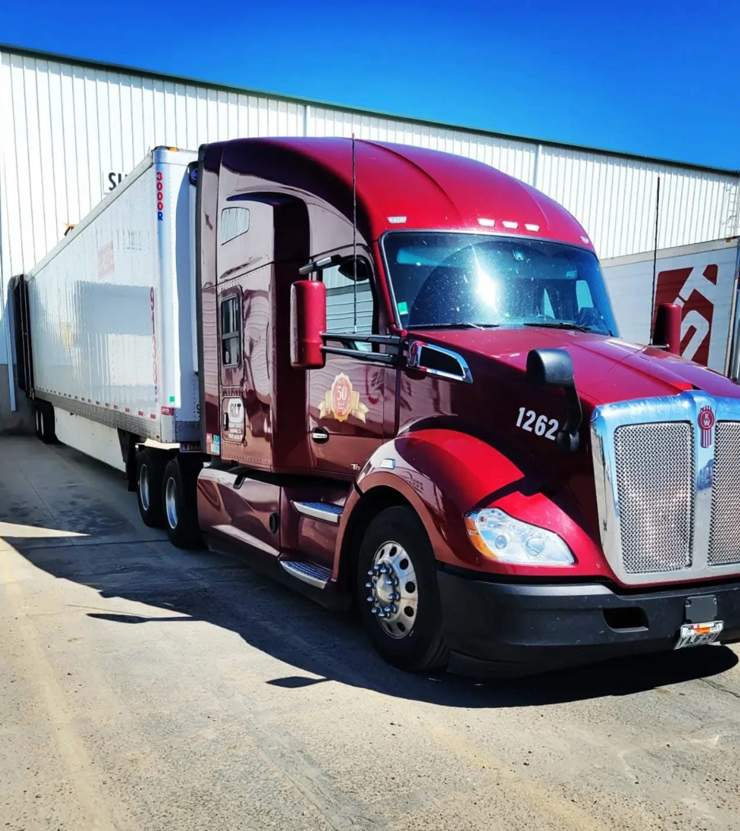 Red semi-truck with a white trailer parked outdoors on a paved surface, with a large white industrial building in the background under a clear blue sky.