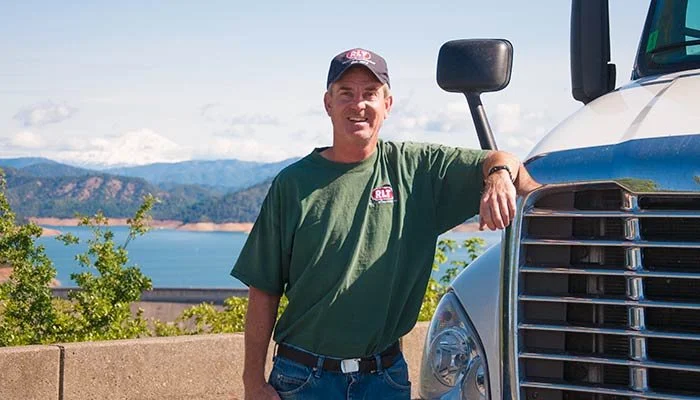 A man wearing a green t-shirt and cap standing next to a large truck with a scenic lake and mountain background.