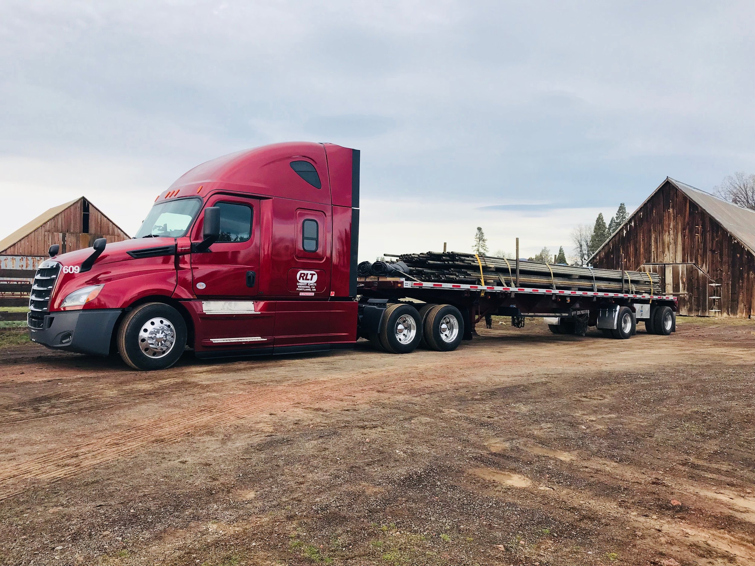 A red semi-truck with a flatbed trailer carrying pipes, parked outdoors with wooden structures and trees in the background.