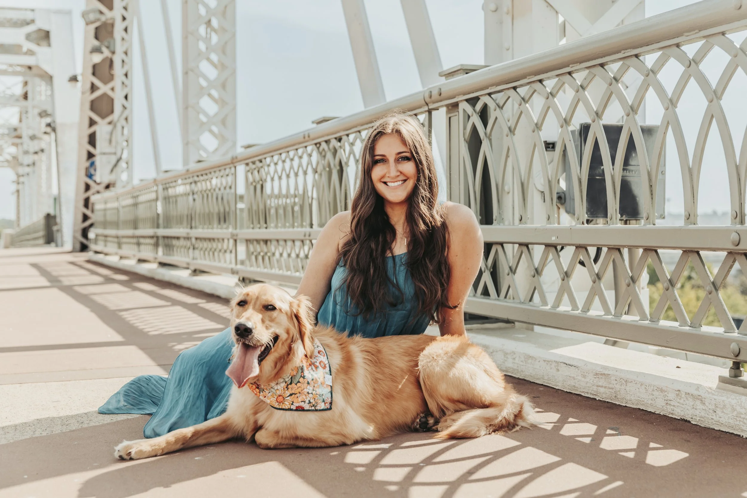 A woman in a blue dress smiling at the camera while lying on the ground next to a golden retriever dog with a floral bandana, on a bridge with metal railings and a clear sky in the background.