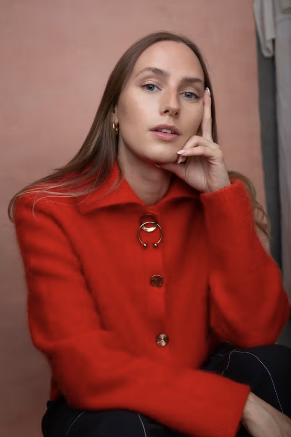 A woman wearing a red jacket with large silver buttons and a decorative circular pin, sitting against a muted pink background, with her hand resting on her face.