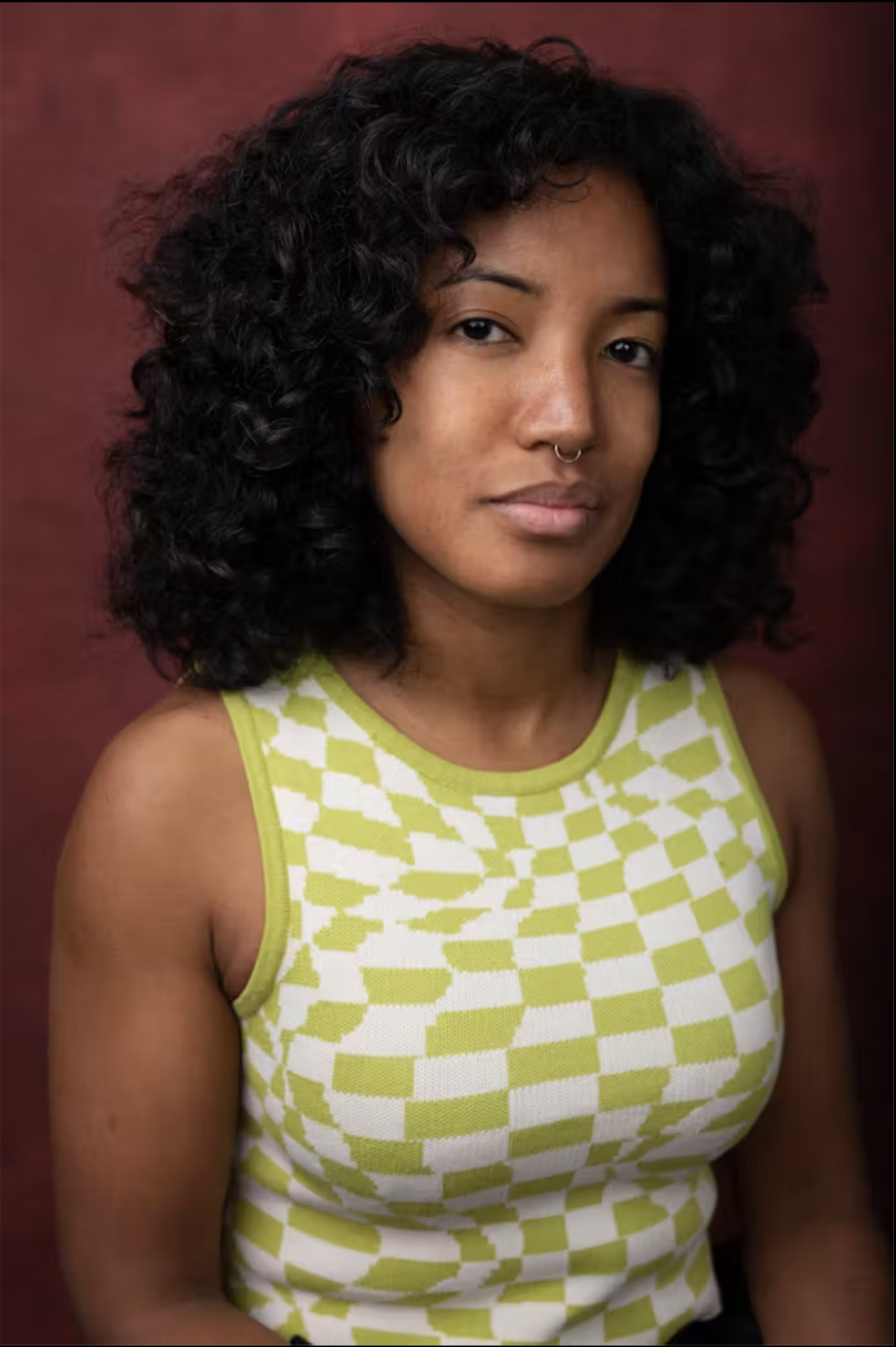 Portrait of a young woman with curly black hair, wearing a yellow and white checkered sleeveless top, against a dark red background.
