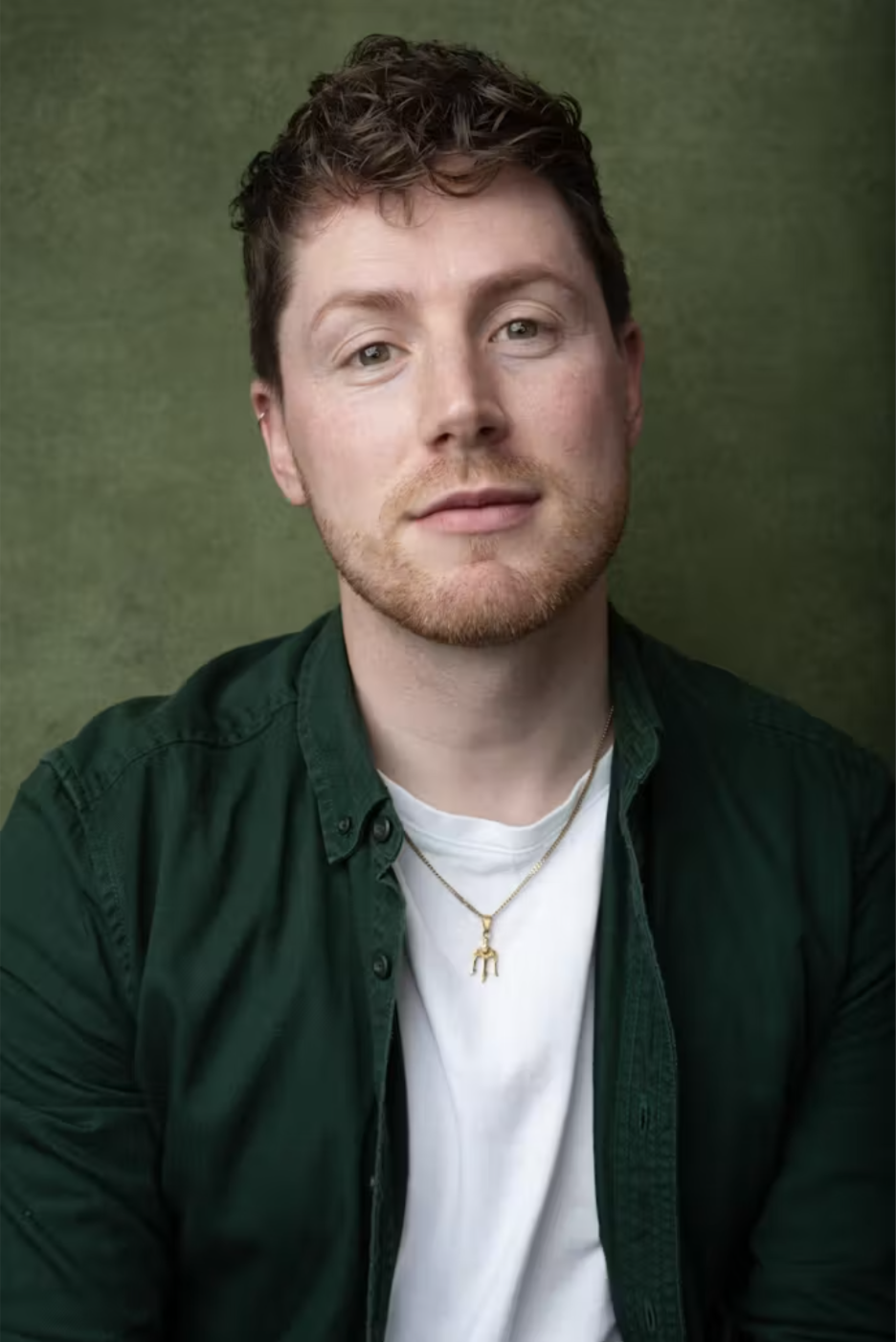 A young man with light skin, short curly brown hair, and a beard, wearing a green button-up shirt over a white t-shirt, with a gold necklace featuring a small figure pendant, against a green background.