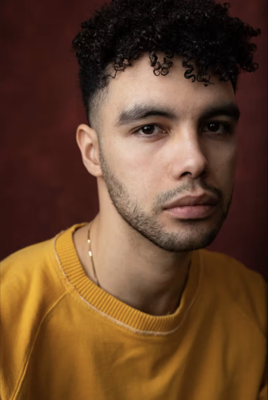 Close-up portrait of a young man with dark, curly hair, light skin, and a trimmed beard, wearing a mustard yellow sweatshirt and a necklace, against a dark red background.