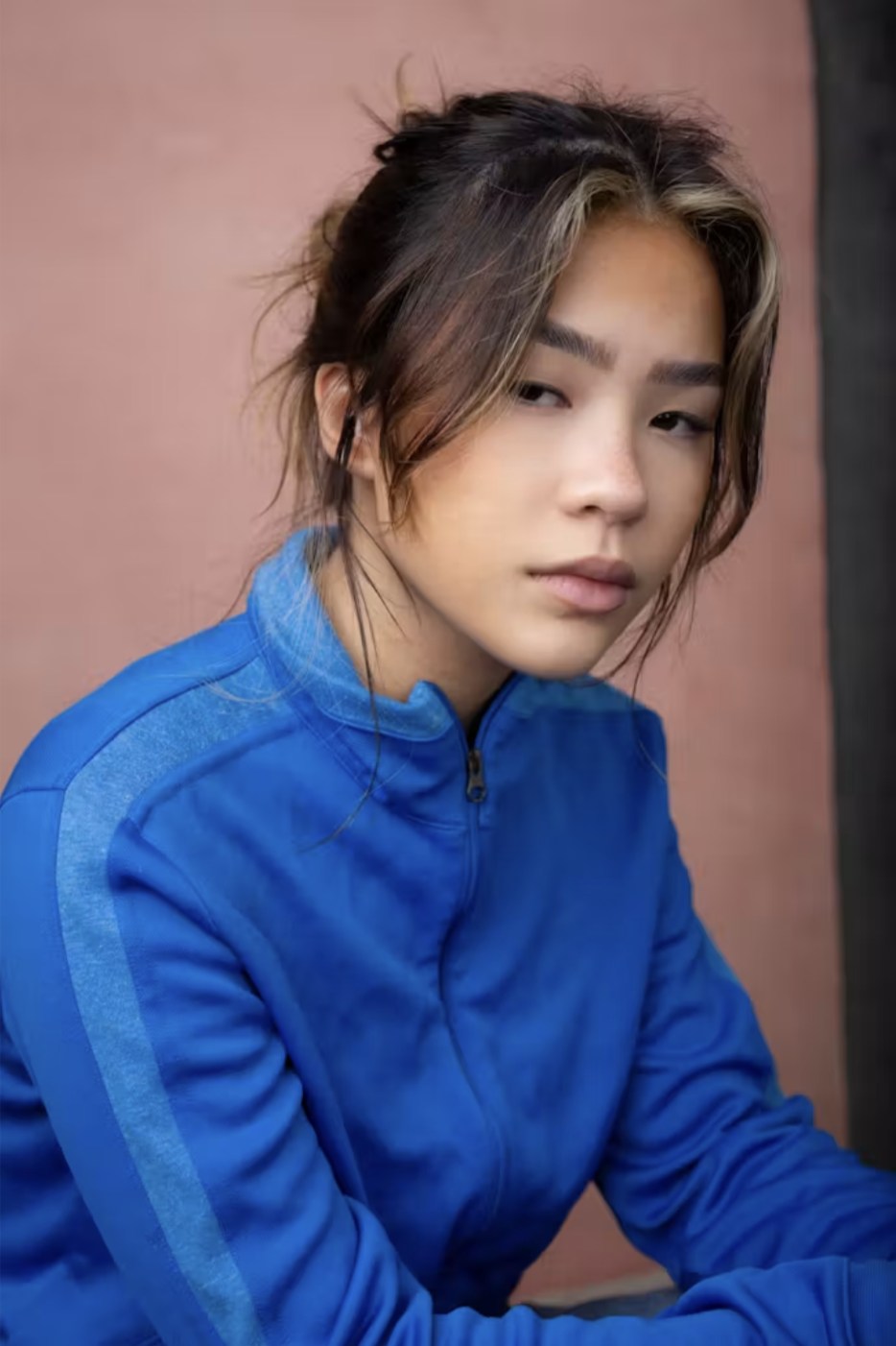 A young woman with tousled dark brown hair with blonde highlights, wearing a blue zip-up jacket with matching stripes on the sleeves, looking at the camera with a neutral expression.