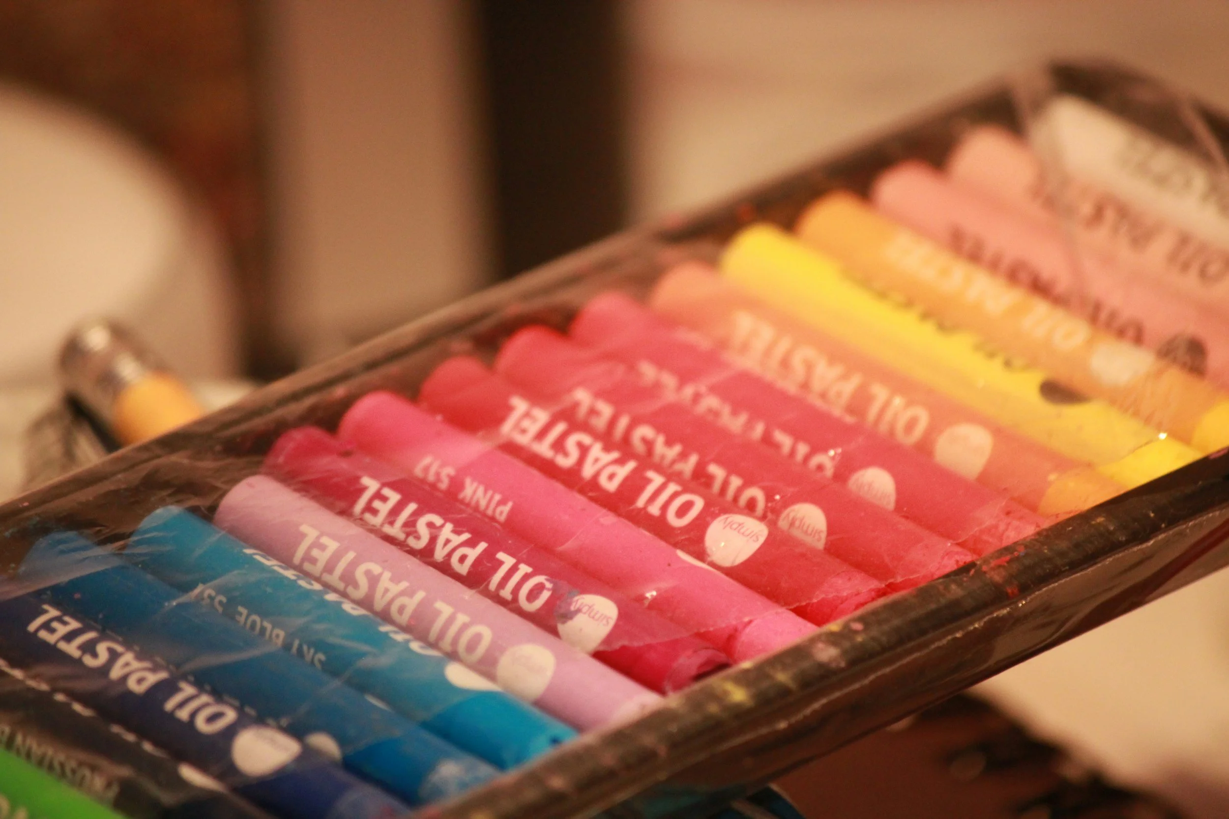 A tray of pastel-colored oil-based paint markers in pink, yellow, orange, blue, and green.