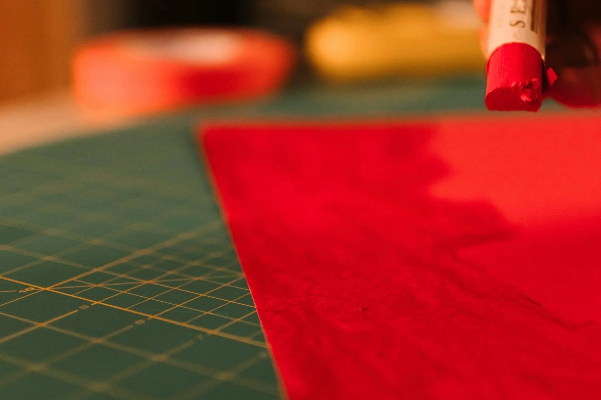 Close-up of a red oil pastel coloring red paper, on a green cutting mat with yellow grid lines.