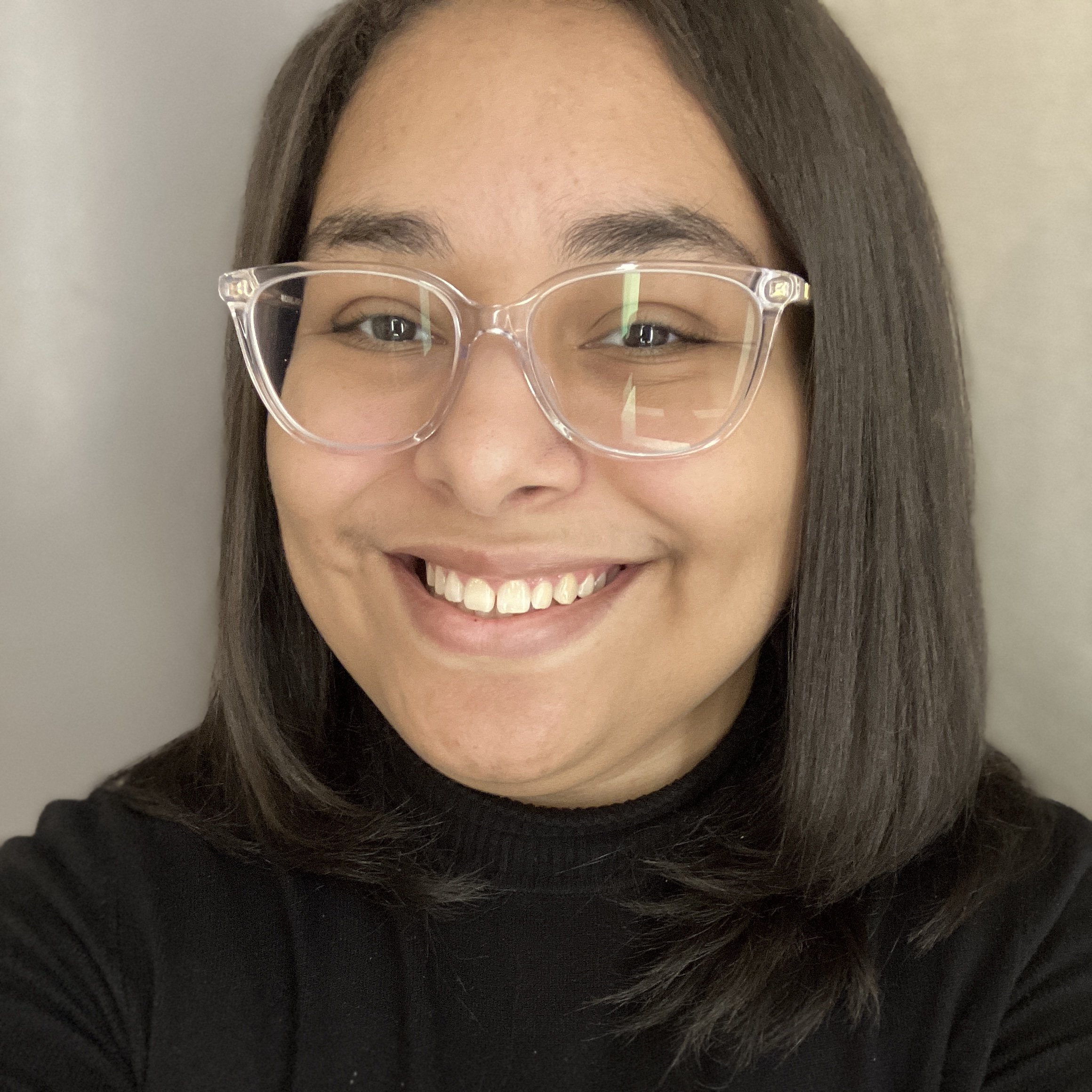 Close-up of a person with dark hair, wearing clear glasses, smiling, with a light-colored background.
