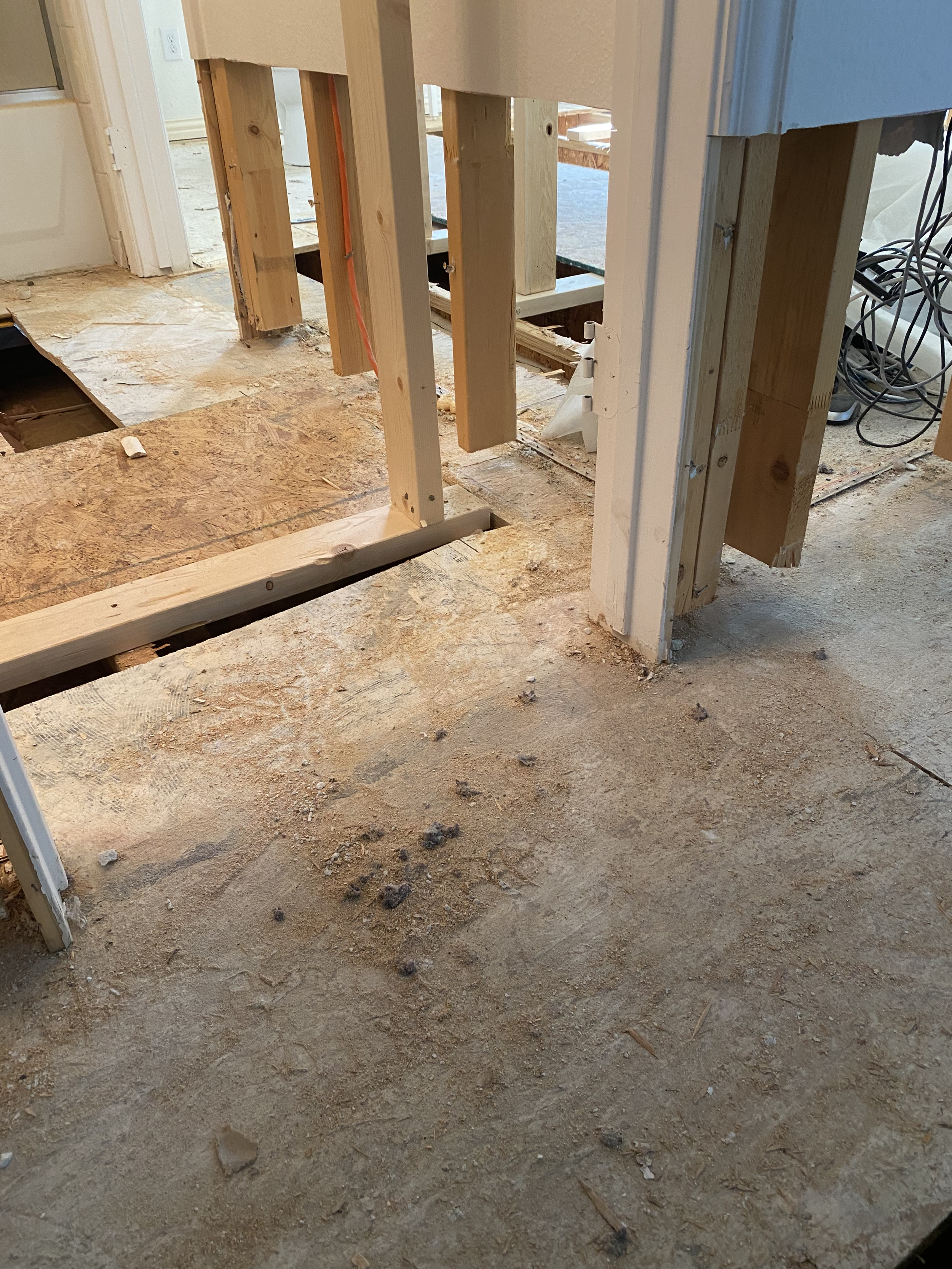 This image shows a room under construction with exposed wooden floor framing, electrical wiring, and unfinished flooring. The floor is made of dirt and debris, and some wooden beams are visible.