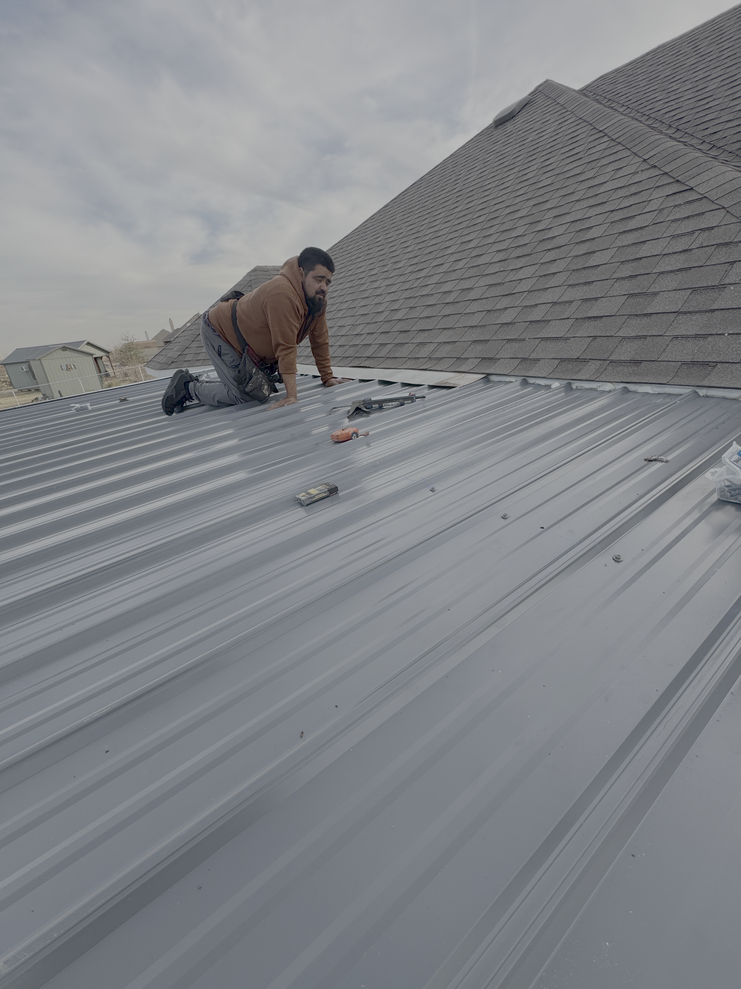 A man working on a metal roof, kneeling and using tools, with a house and cloudy sky in the background.