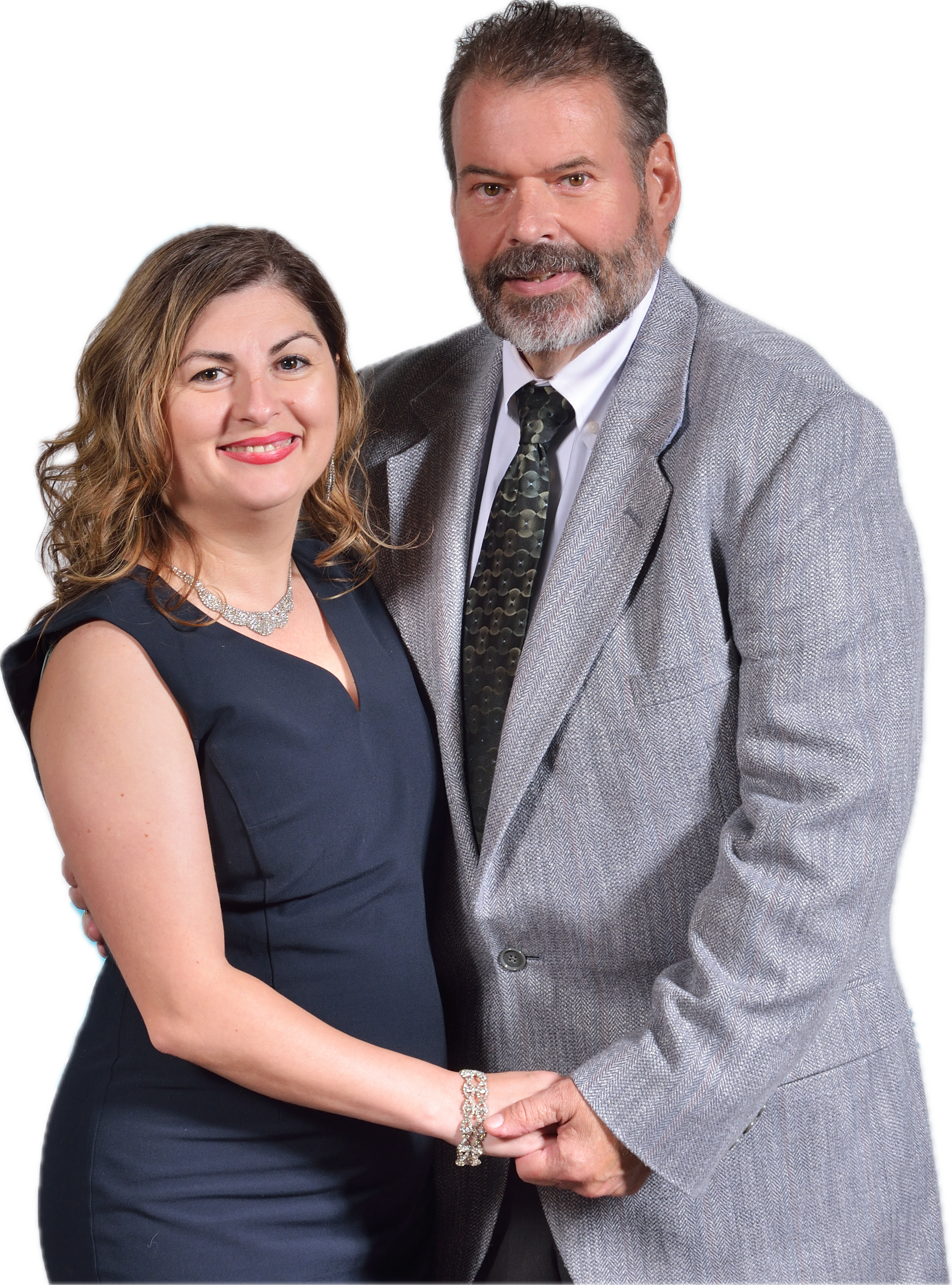 A man and woman dressed in formal attire are standing close together, holding hands and smiling for a portrait. The woman has long, wavy light brown hair, and is wearing a dark blue dress with a necklace and bracelet. The man has short, gray hair and a beard, and is wearing a light gray blazer, a white shirt, and a patterned tie.