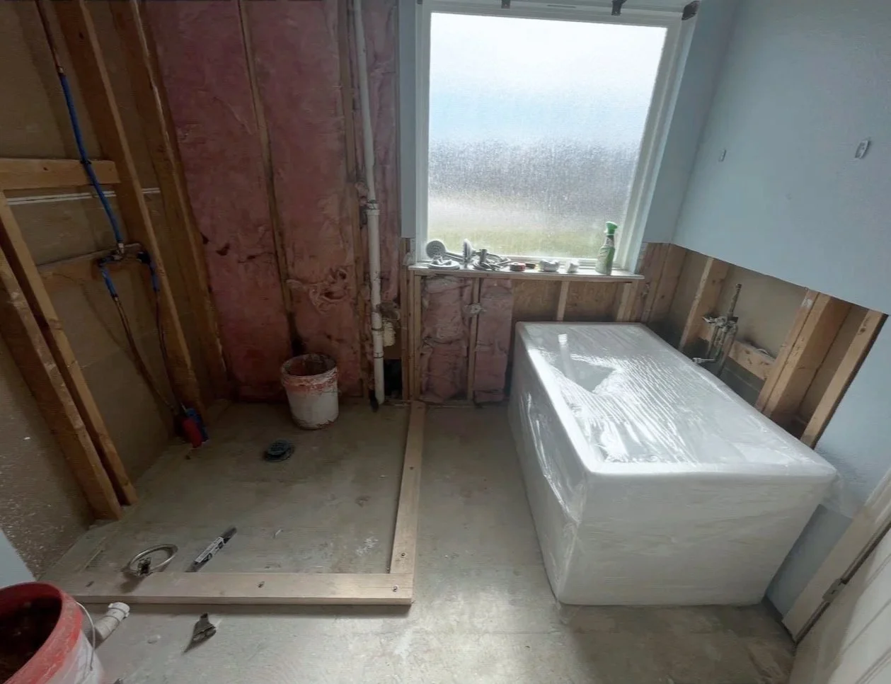 Bathroom under construction with exposed wooden framing, insulation, tub wrapped in plastic, and window with a seagull in the background.