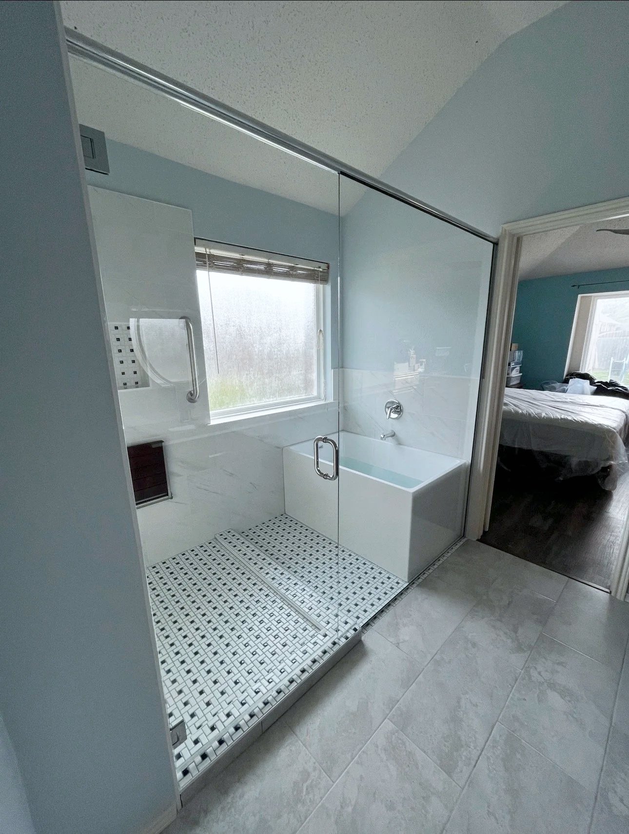 Modern bathroom with a glass-enclosed shower, white bathtub, and window, adjacent to a bedroom with a bed and window.