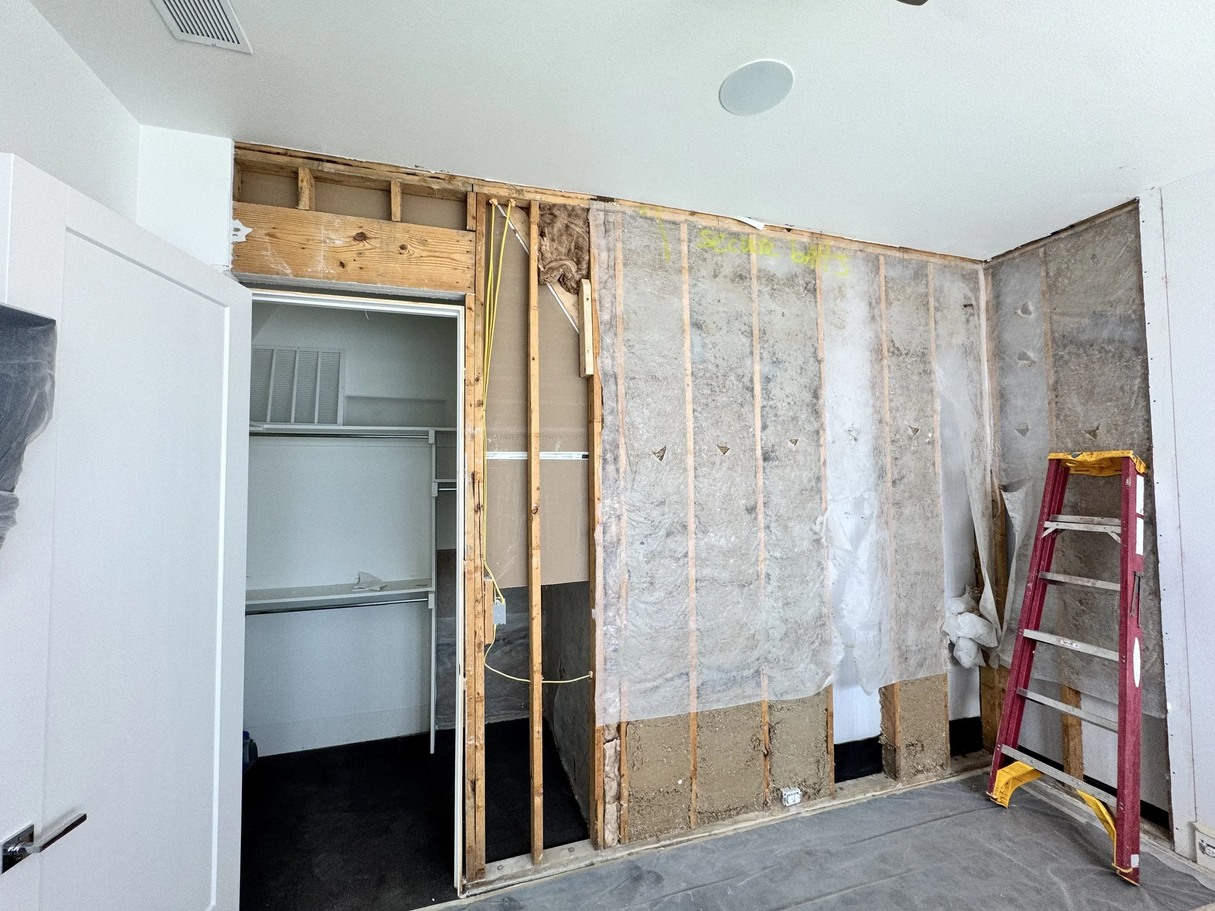 Interior of a room under renovation with walls partially stripped, exposing wooden studs and insulation. A red ladder is on the right side, and a doorway with white frames leading to another room.