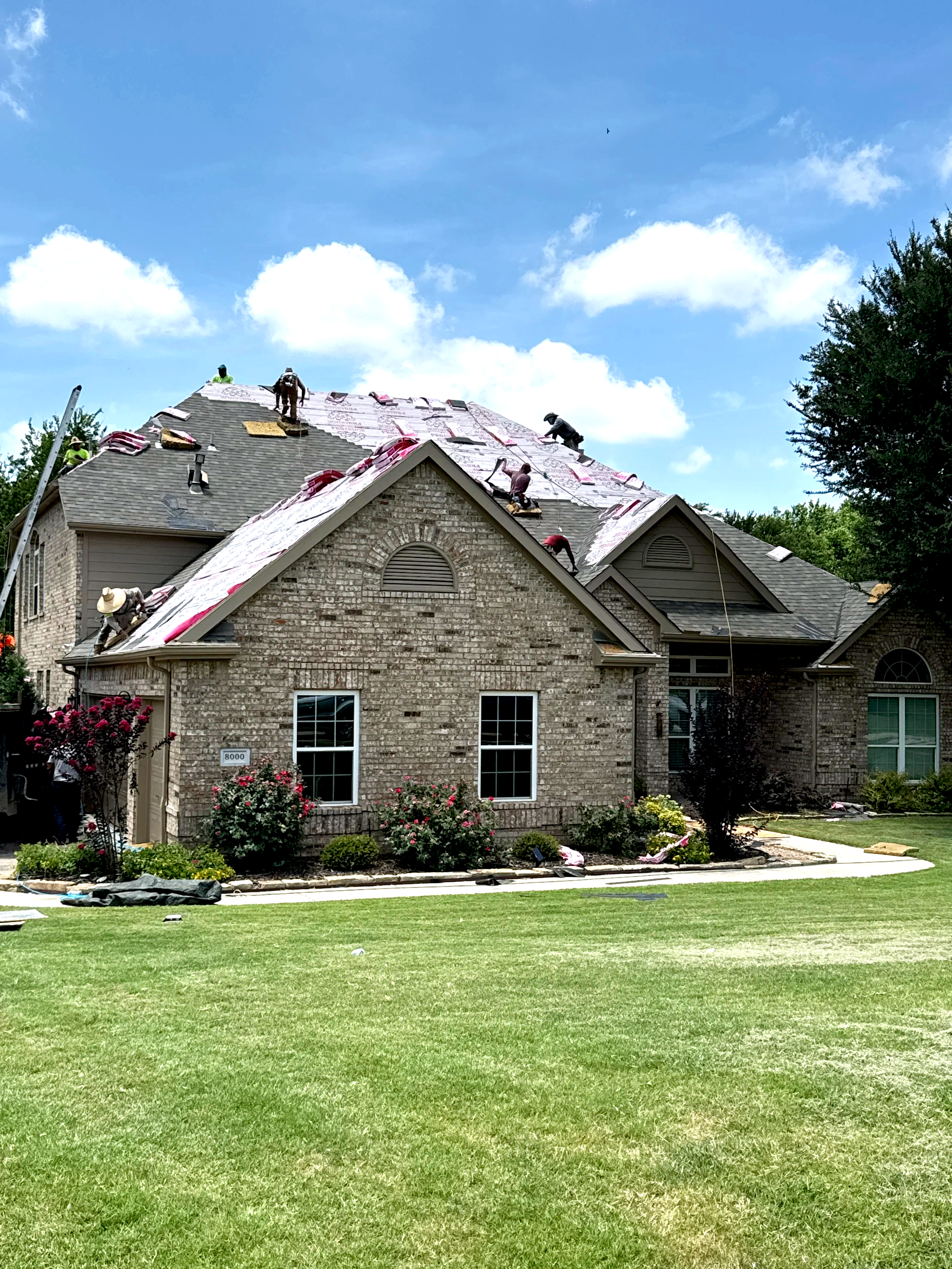 Workers installing new roofing on a brick house with a large front yard and well-maintained lawn, under a blue sky with some clouds.