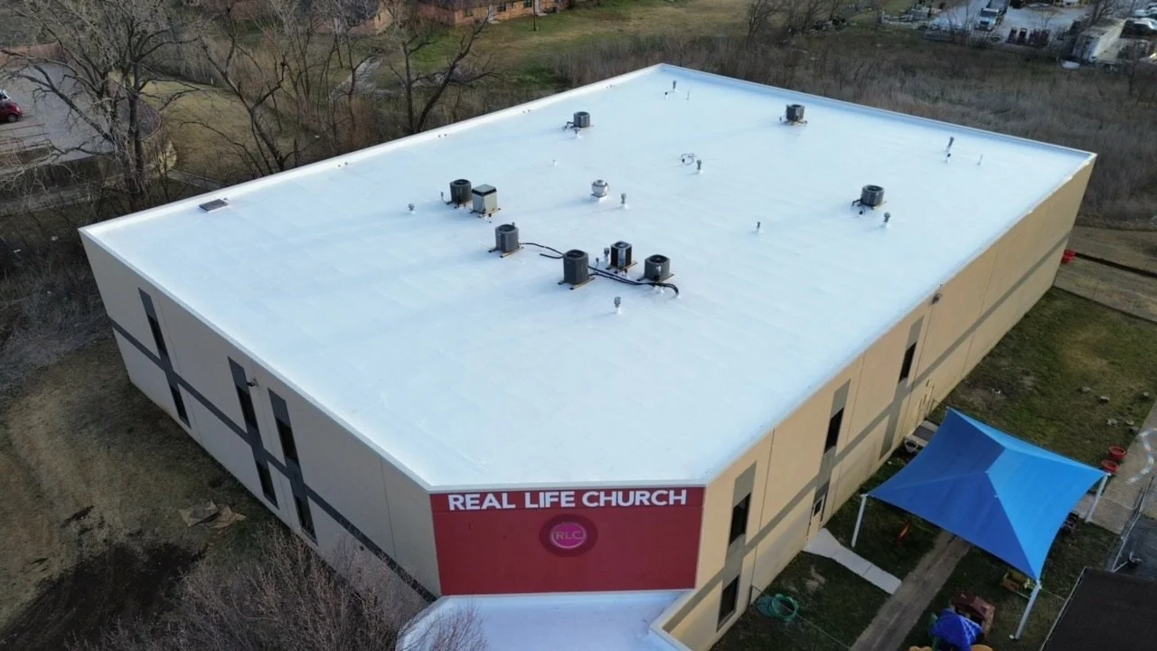 Aerial view of a beige rectangular building with a flat white roof, labeled 'Real Life Church,' surrounded by a small yard with a blue canopy and pathways.