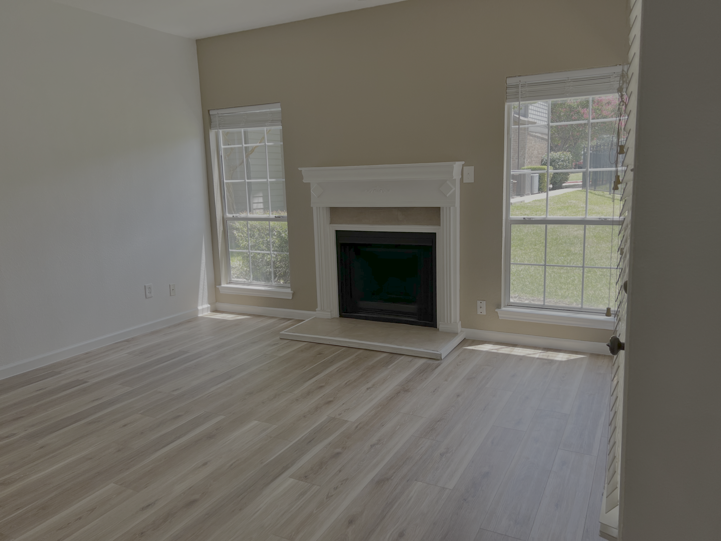 Interior view of a living room featuring a fireplace mantel, large windows with blinds, and light-colored wood flooring.