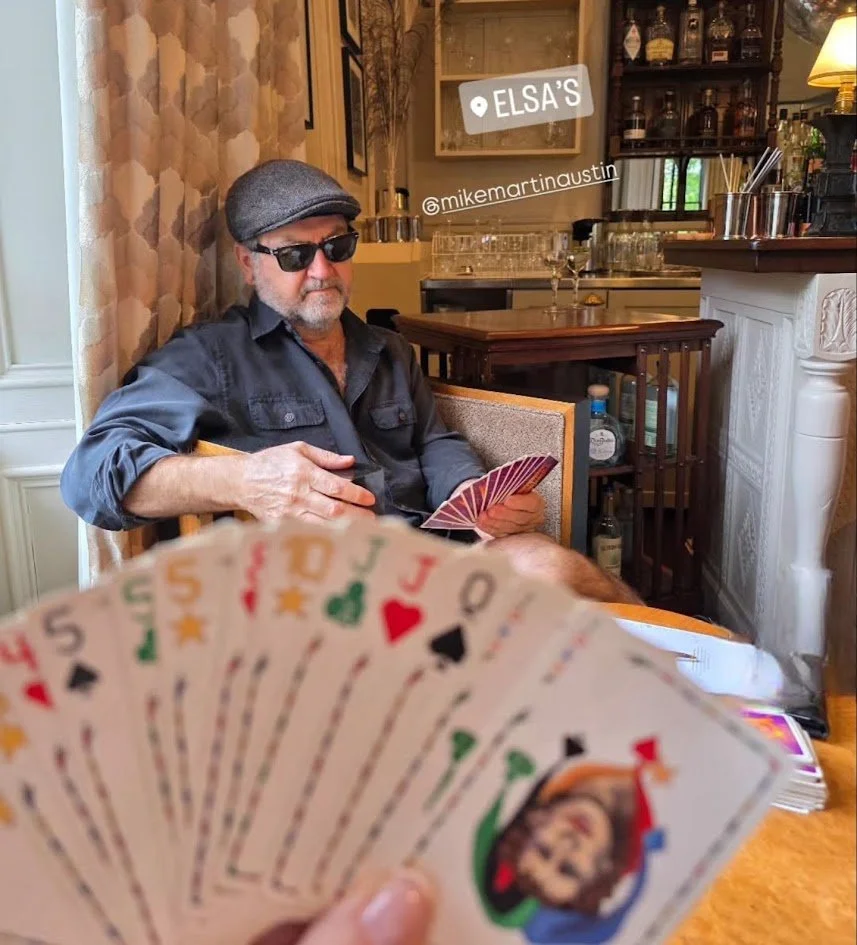 A man sitting in a cozy, warmly lit restaurant or bar, wearing sunglasses and a gray flat cap, holding a hand of playing cards.