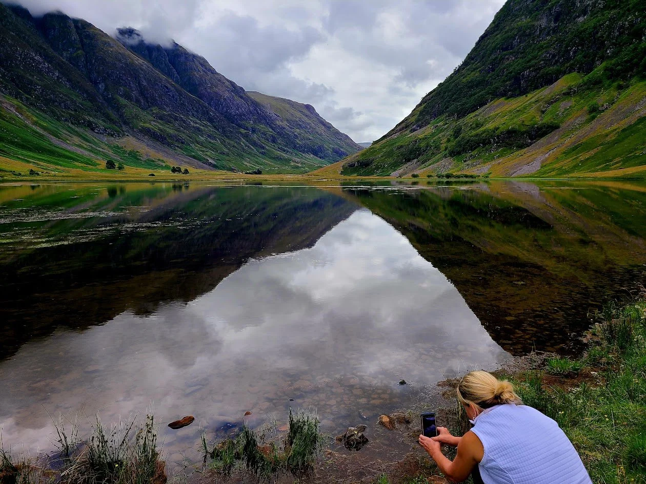 A woman with blonde hair in the mountains, by a lake taking a photo, Glencoe Scotland