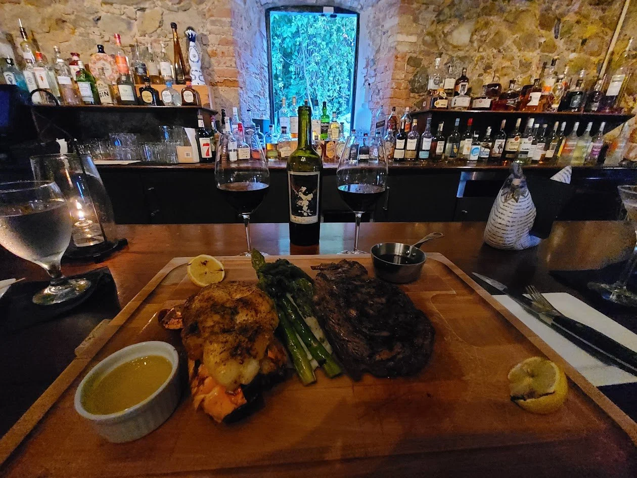 A wooden table with a plating of grilled steak, roasted vegetables, green onions, lemon wedges, and dipping sauces. Two glasses of red wine and a bottle are centered. Background features a bar with various bottles and glasses, and a window with blue lighting in a rustic stone-walled room.