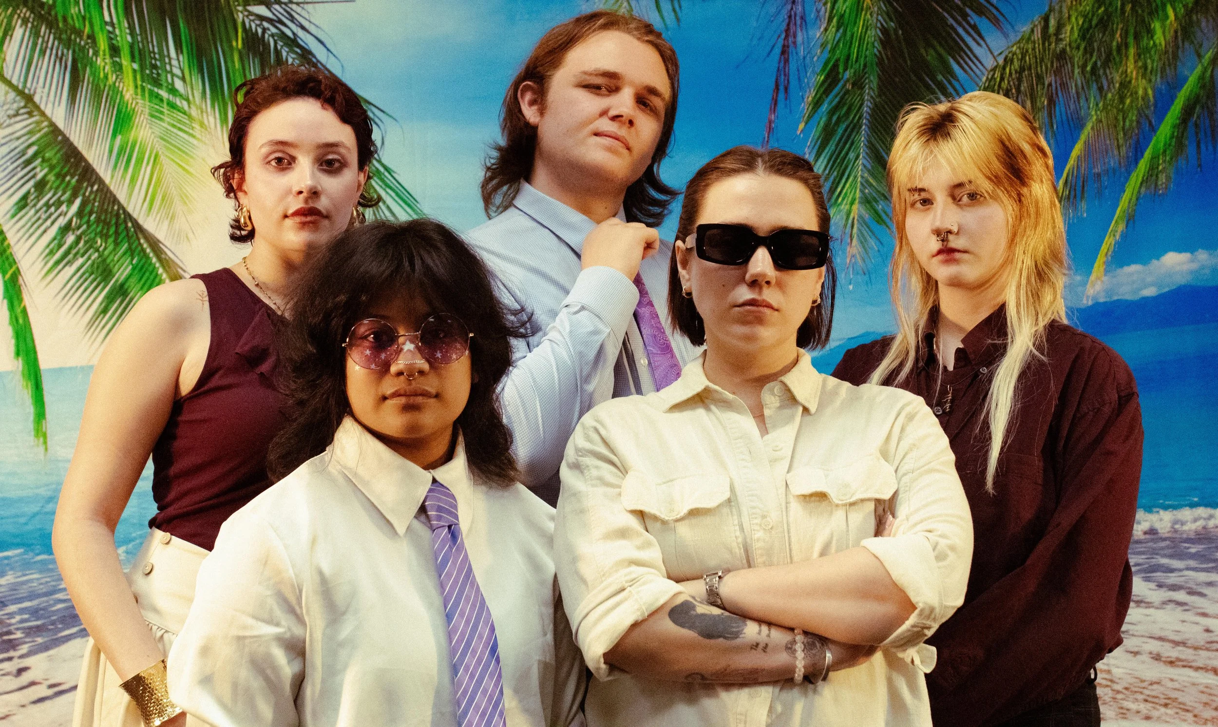 A group of six diverse young adults posing in front of a tropical beach background with palm trees and blue sky.