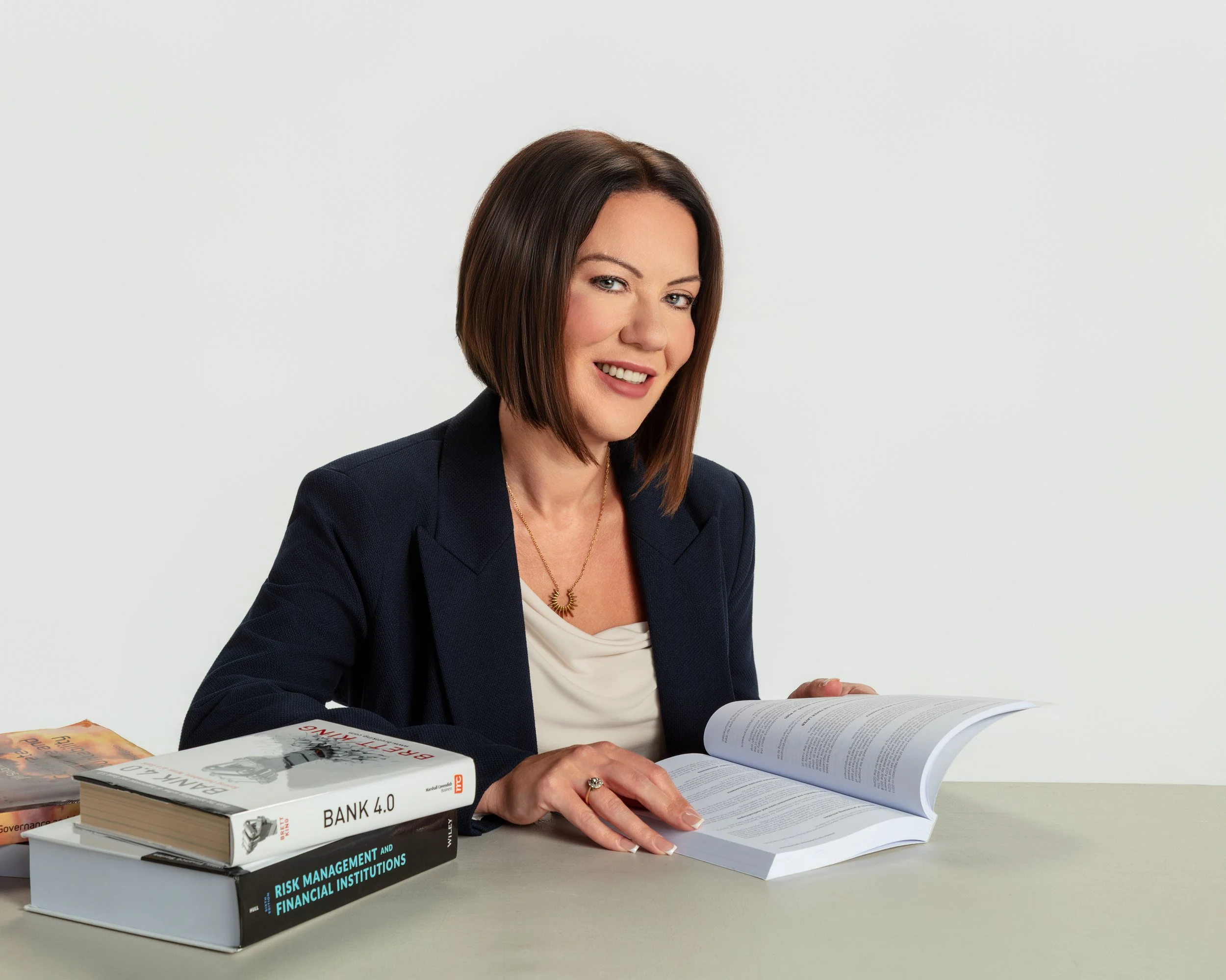 A woman with shoulder-length brown hair and blue eyes is sitting at a table with an open book and several stacked books about banking, risk management, and financial institutions. She is wearing a dark blazer, a white shirt, and a gold necklace, smiling at the camera.