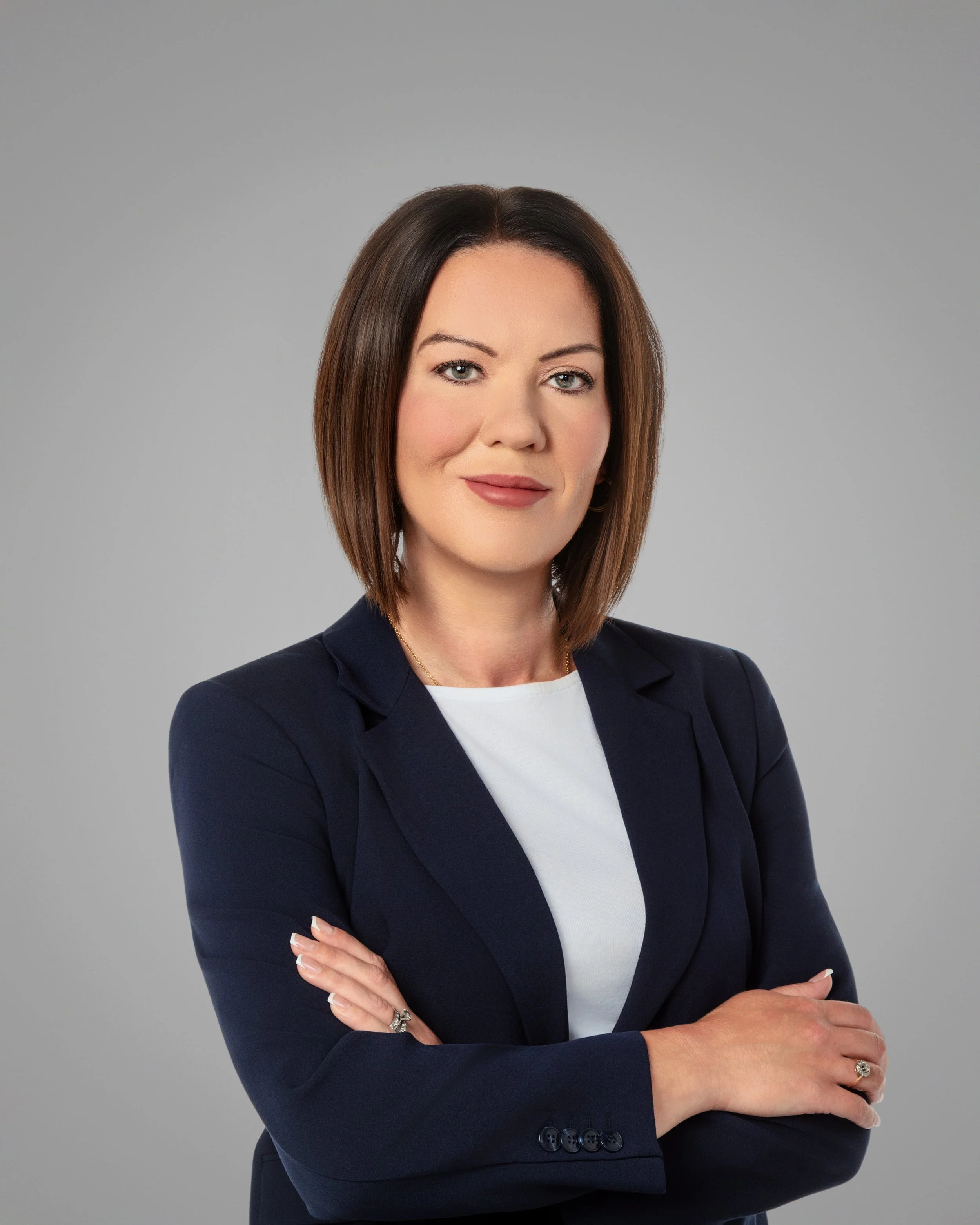 Professional woman with shoulder-length brown hair, light blue eyes, wearing a navy blazer, white shirt, and a ring on her ring finger, posing with crossed arms against a gray background.