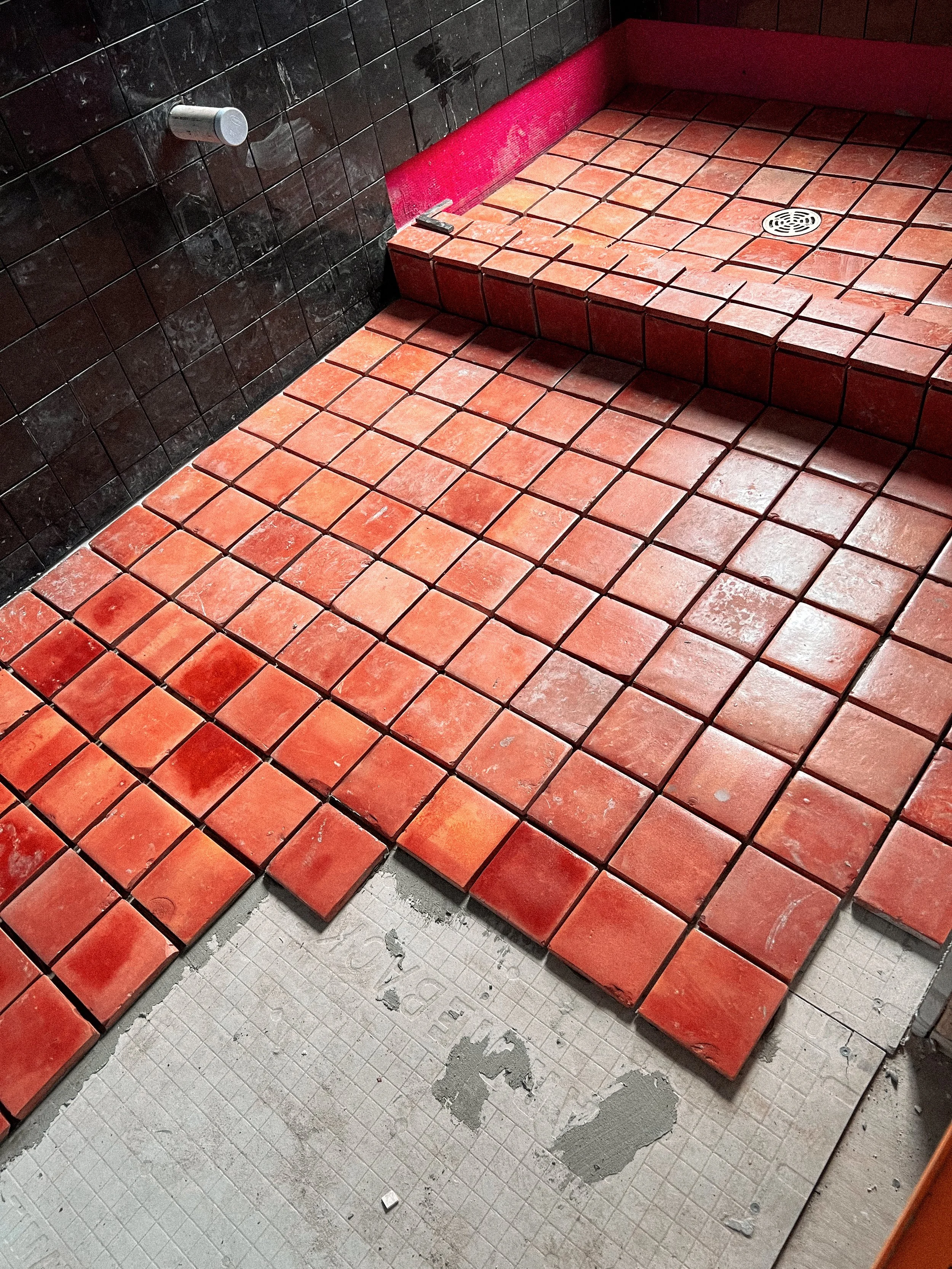 The image shows red and black tiled flooring likely in a bathroom or shower area. The shower area has a drain on the floor and a ledge with a small tool or object on it. The black tiles are on the wall, while the floor and shower ledge are covered with red tiles.