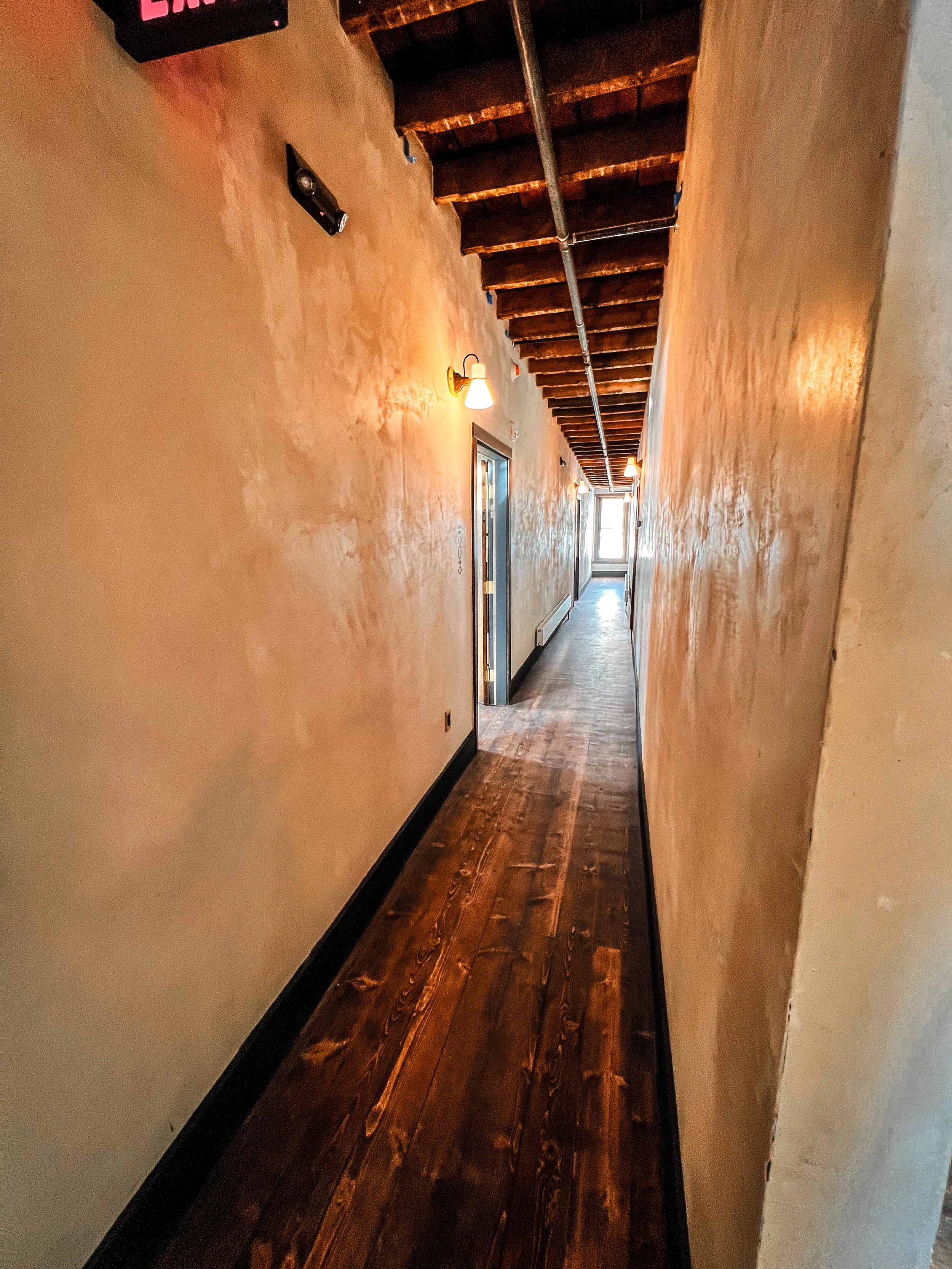 Long narrow corridor with wooden floors, textured beige walls, exposed wooden ceiling beams, and wall-mounted lamps providing warm lighting. There are ceiling pipes and a door at the end of the hallway.