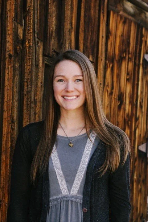A young woman with long brown hair and a bright smile stands in front of a wooden wall, wearing a gray top and black jacket.