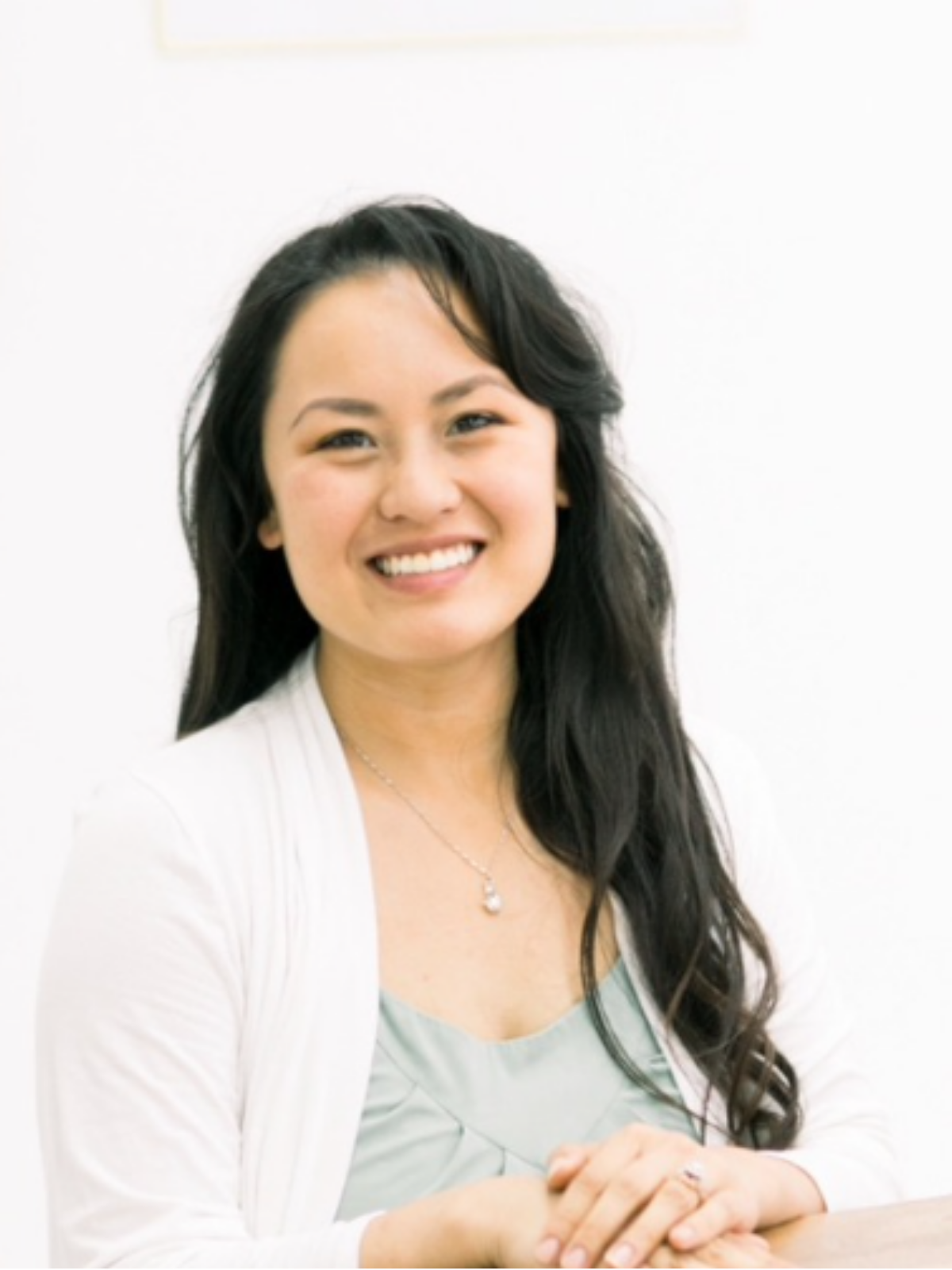 A smiling woman with long black wavy hair, wearing a white blazer over a light-colored top, and a necklace, seated with hands clasped in front of her against a plain white background.