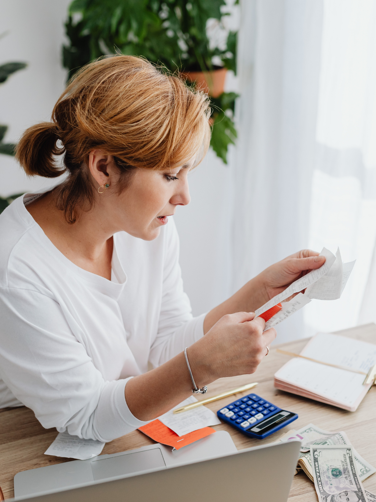 A woman with short red hair reads a receipt at a cluttered desk with receipts, a calculator, a laptop, and money.