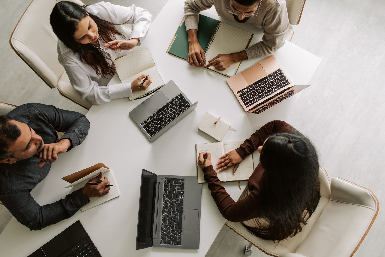 Top view of four people sitting around a white table working on laptops, taking notes, and discussing, with notebooks and pens.