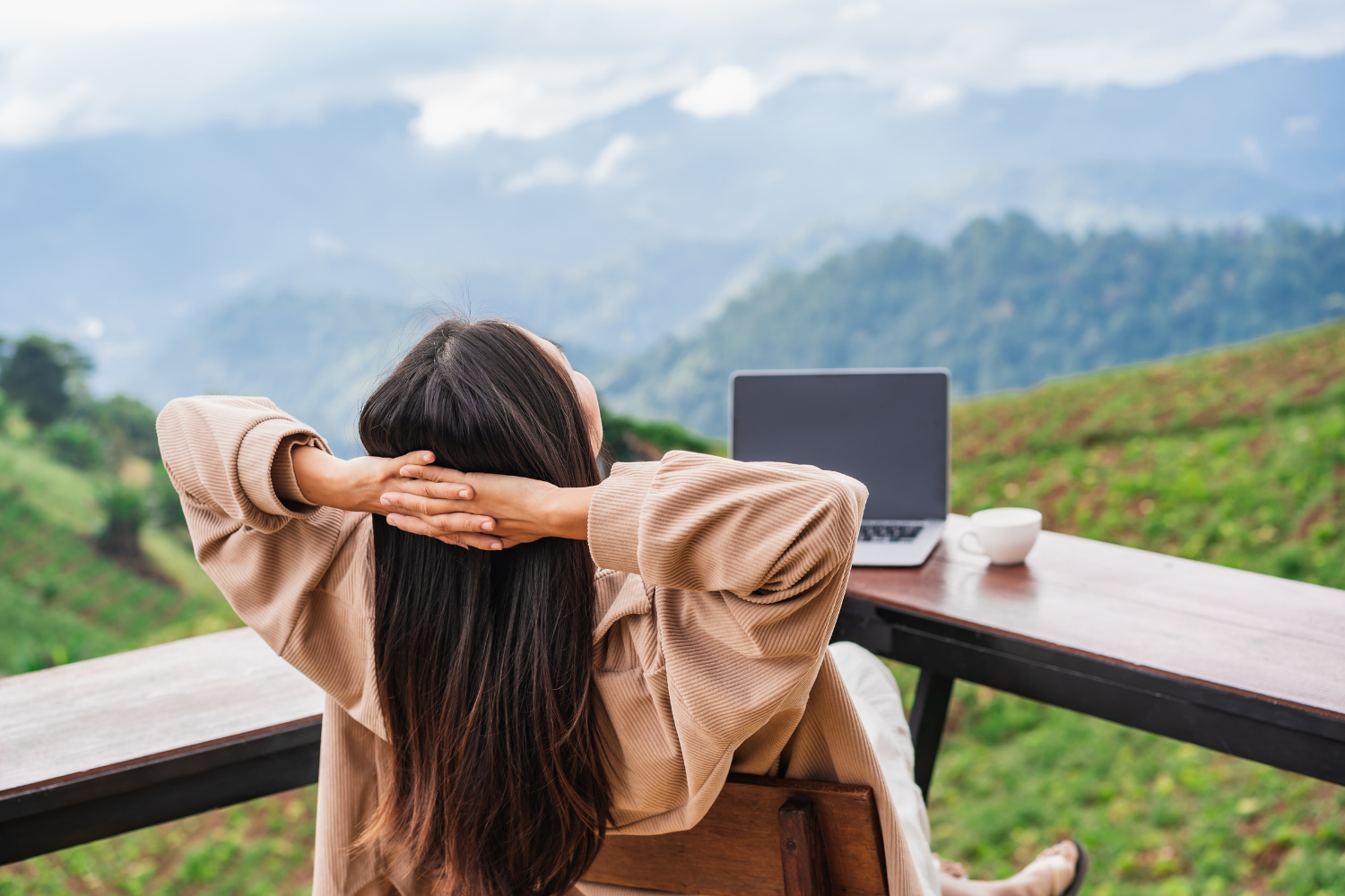 Woman relaxing at a wooden table outdoors with a laptop and coffee cup, overlooking lush green hills and mountains in the distance.