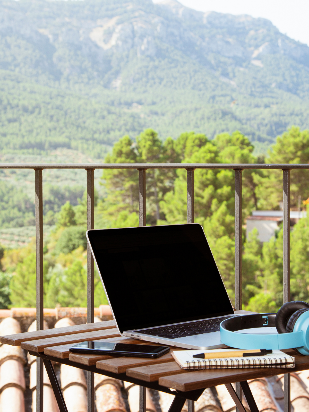 A laptop, smartphone, notebook, pen, headsets, and a tablet on a wooden table on a balcony with a mountain view and green trees in the background.