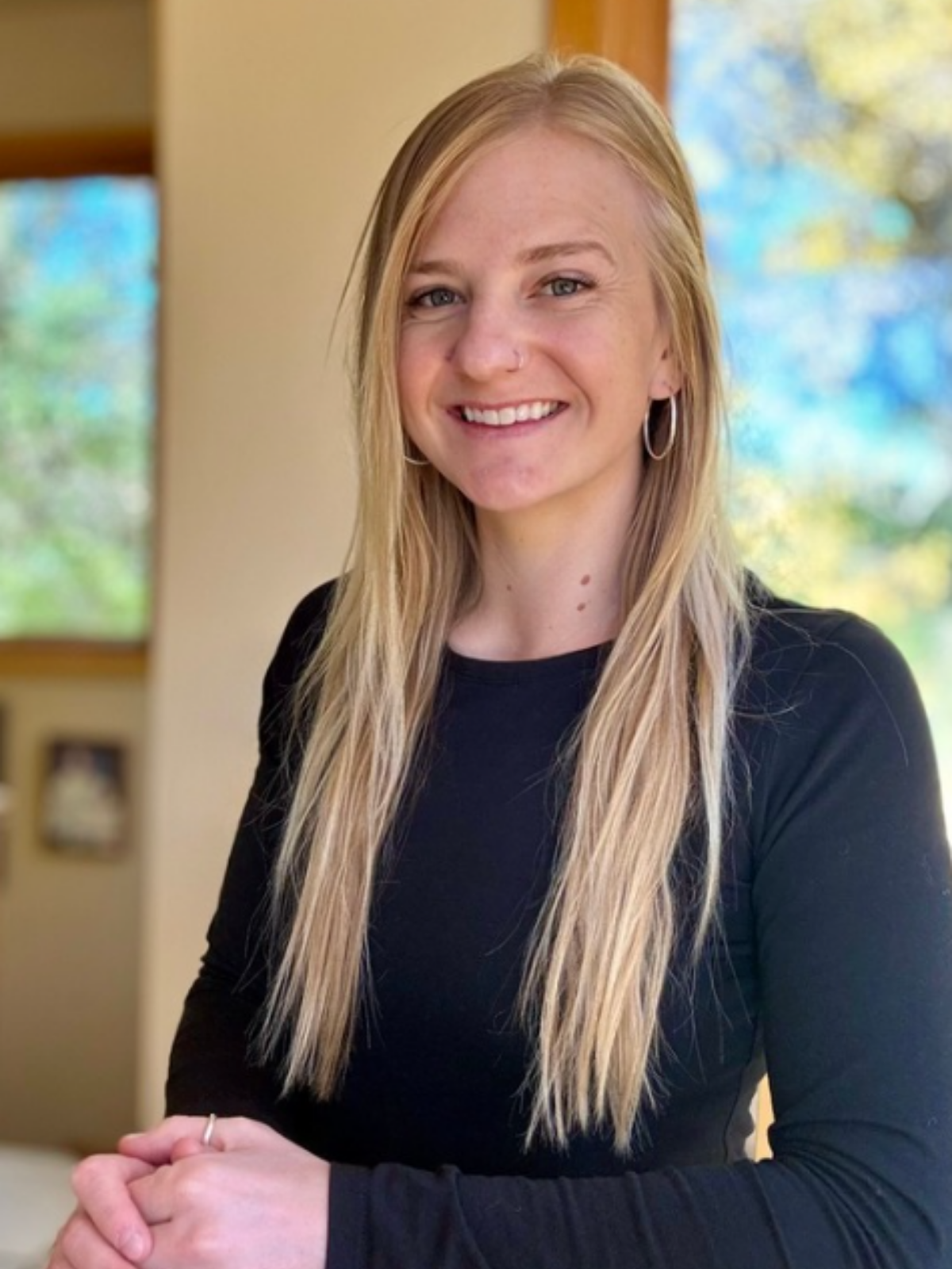 A smiling woman with long blonde hair, wearing hoop earrings and a black shirt, standing indoors with a window showing a blurred outdoor scene in the background.