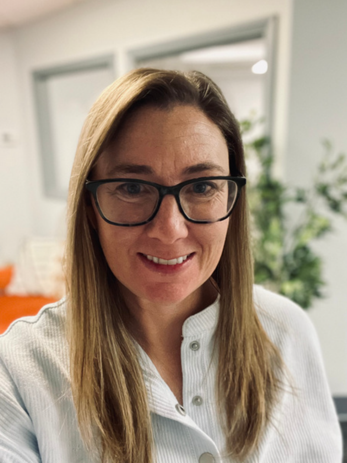 A woman with long blonde hair, black glasses, and a white shirt smiling in an office setting.