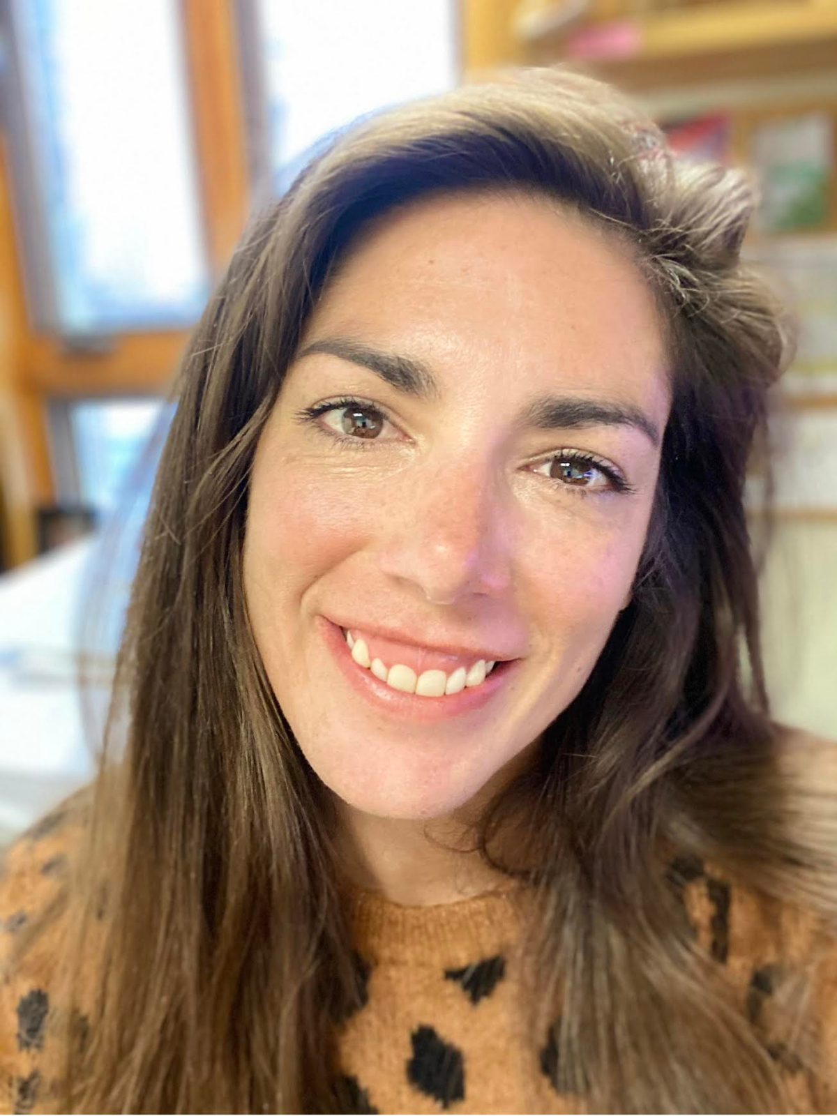 A smiling woman with long brown hair and brown eyes, wearing a brown sweater with black patterns, indoors with a blurry background of books and windows.