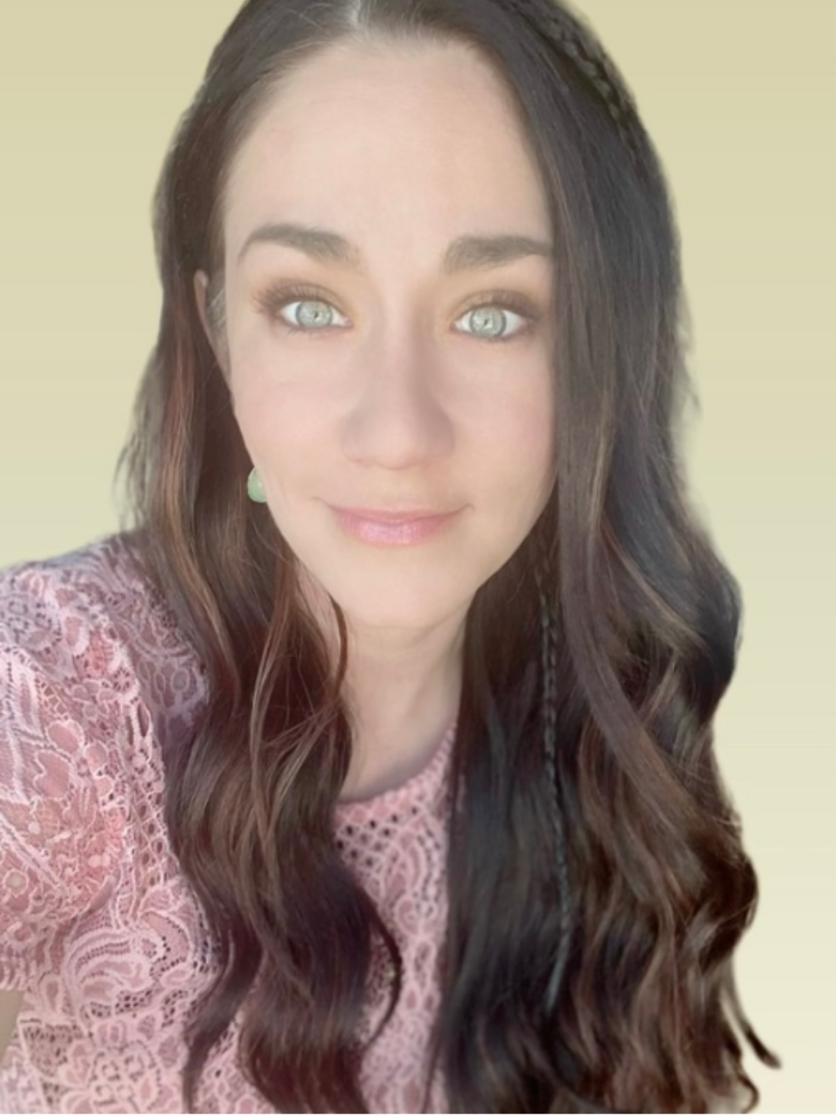 A young woman with long dark wavy hair, light eyes, and light skin, wearing a pink lace top and light green earrings, smiling against a pale yellow background.