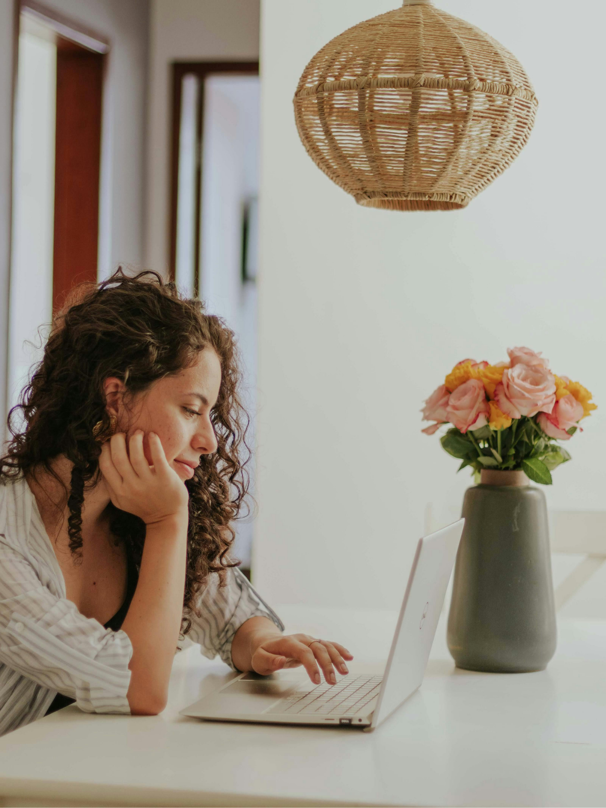 A woman with curly hair sitting at a white table, working on a laptop with a focused expression, in a room with a vase of pink and yellow flowers and a wicker lampshade overhead.