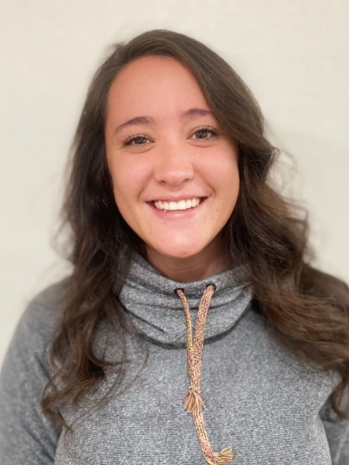 A young woman with long wavy brown hair, smiling, wearing a gray hoodie with a drawstring, standing against a plain light-colored wall.