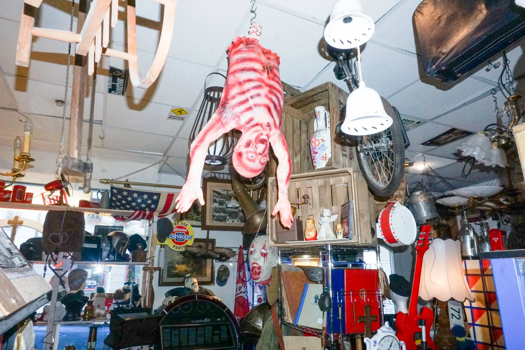 Ein Laden mit verschiedenen Vintage- und Antikobjekten, darunter eine große rot-weiße Dekofigur eines Tieres, eine Stahlfeder, Kerzenständer, ein Wasserkocher, eine Fahrradfelge, eine Matrosenmütze und andere dekorative Gegenstände, alles an der Deck